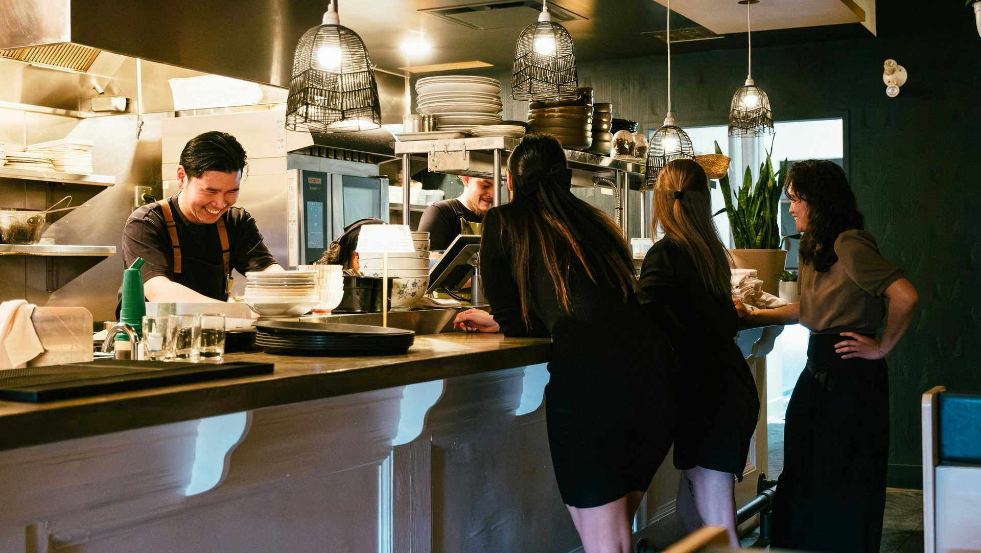 A restaurant counter with people talking to a smiling chef. Overhead lights hang above.