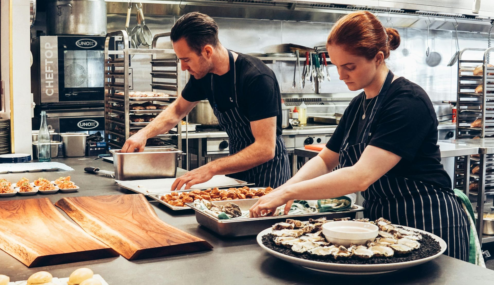 Two chefs in black shirts and aprons preparing food in a commercial kitchen.