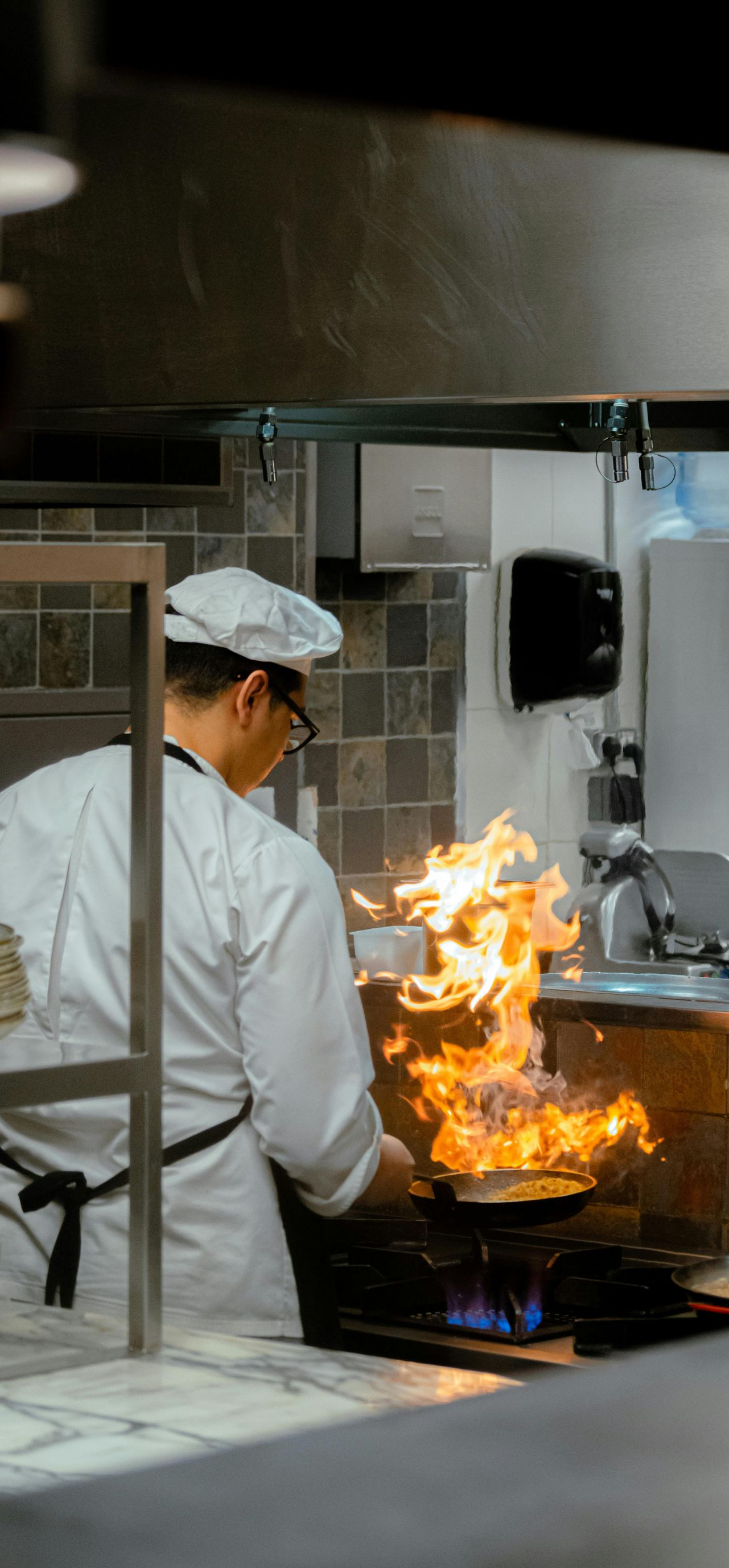 Chef in white coat, hat, and glasses flambéing food in a pan over a stove with large flames.