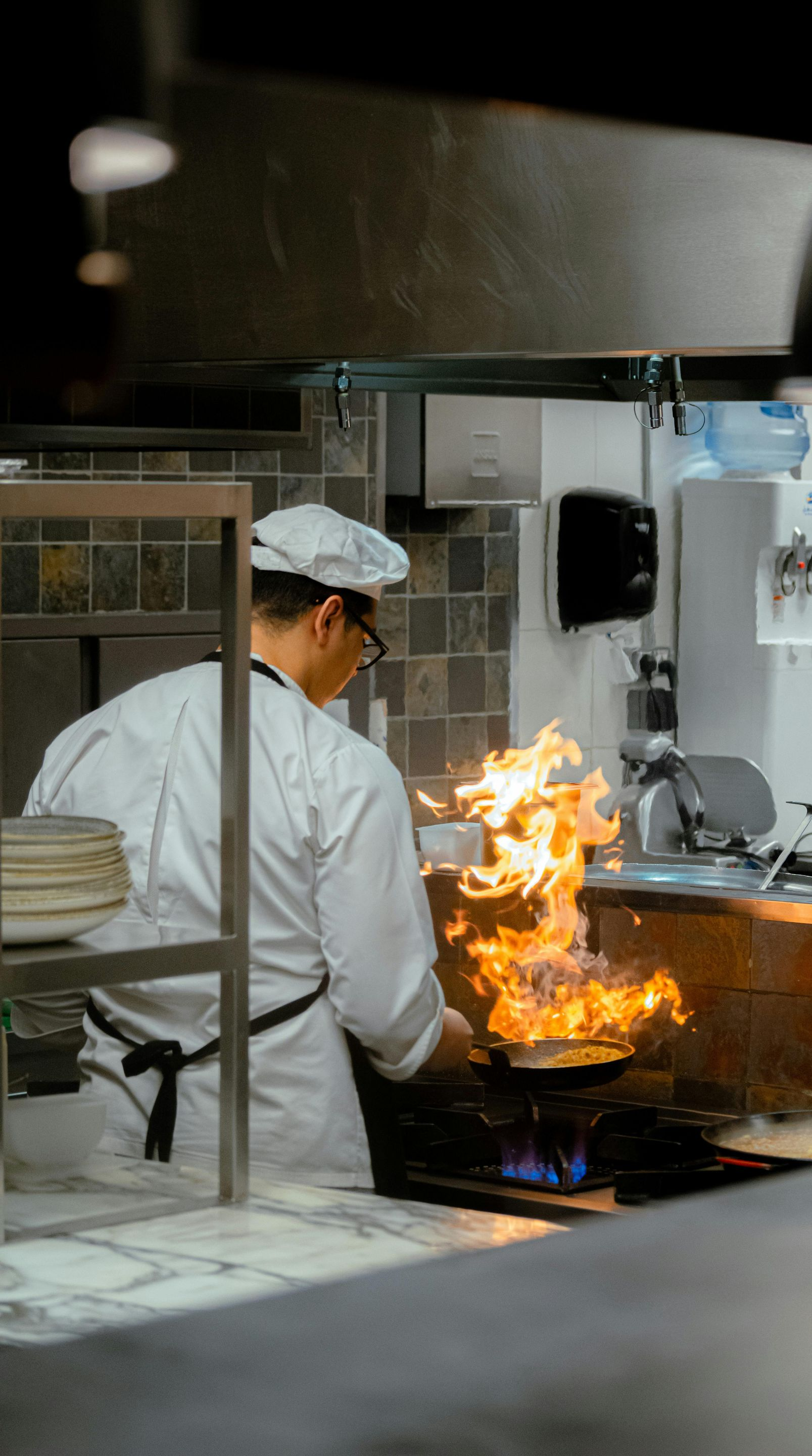 Chef in white uniform flips food in a pan with flames, commercial kitchen setting.