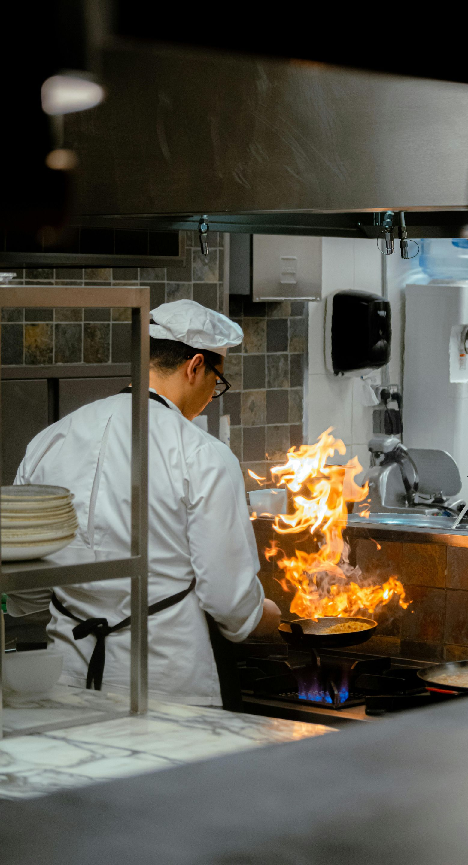 Chef in white uniform flips food in a pan with flames, commercial kitchen setting.
