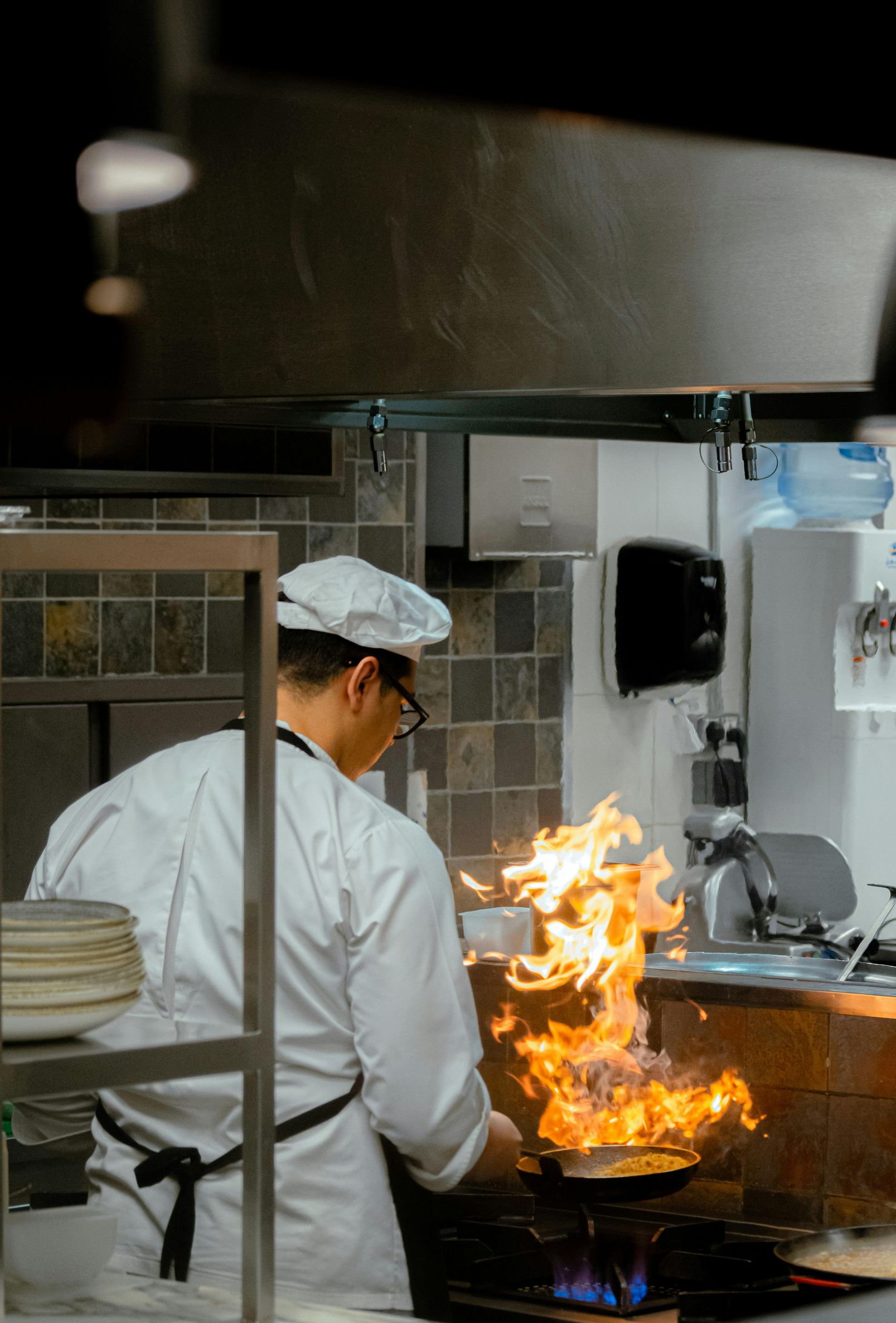 Chef in white uniform flips food in a pan with flames, commercial kitchen setting.