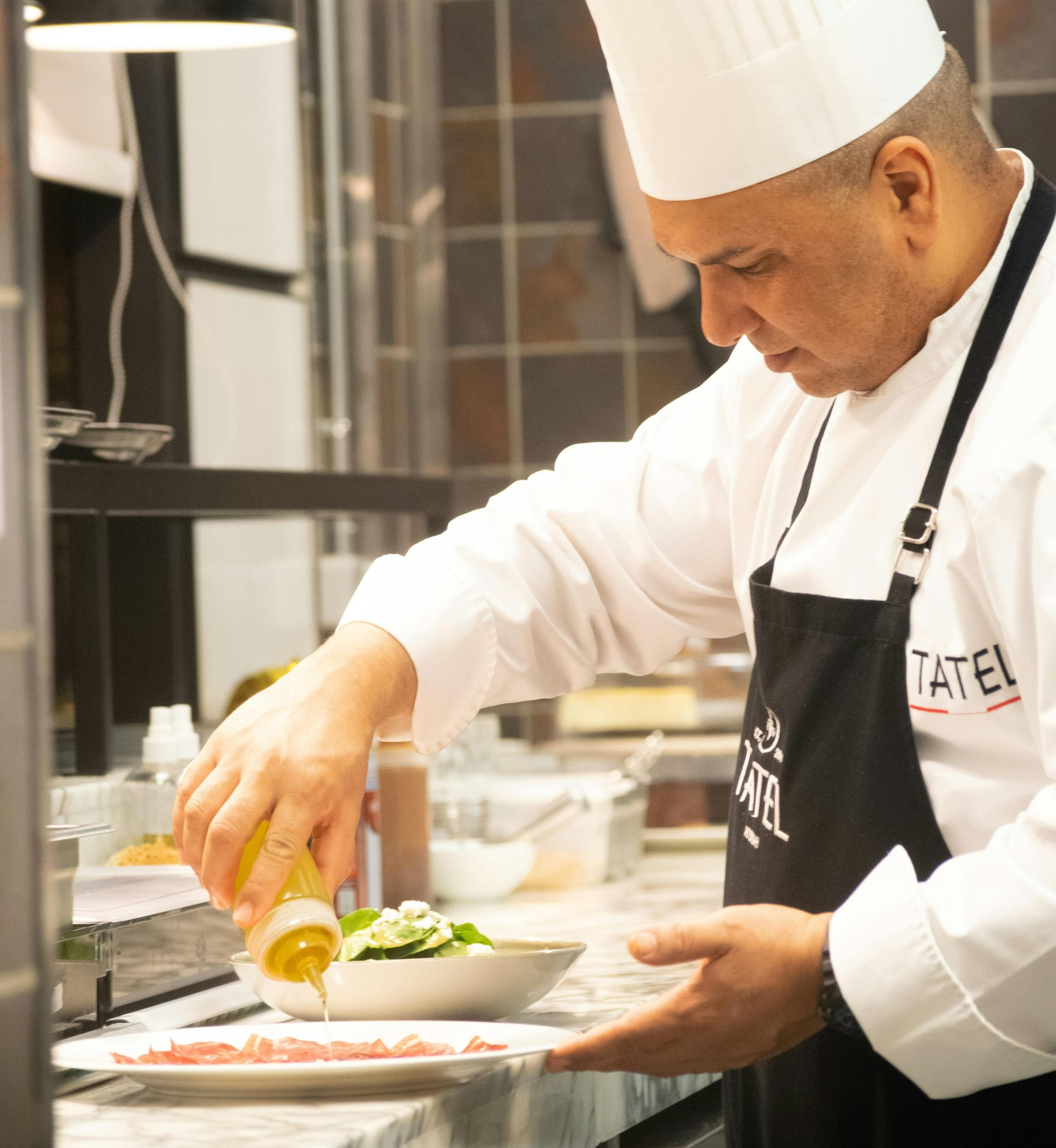 Chef in a white uniform adding sauce to a plate of food in a kitchen.