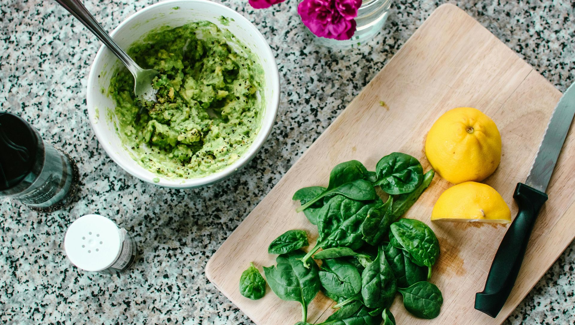 A white bowl of guacamole next to a cutting board with spinach, a cut lemon, and a knife on a speckled countertop.