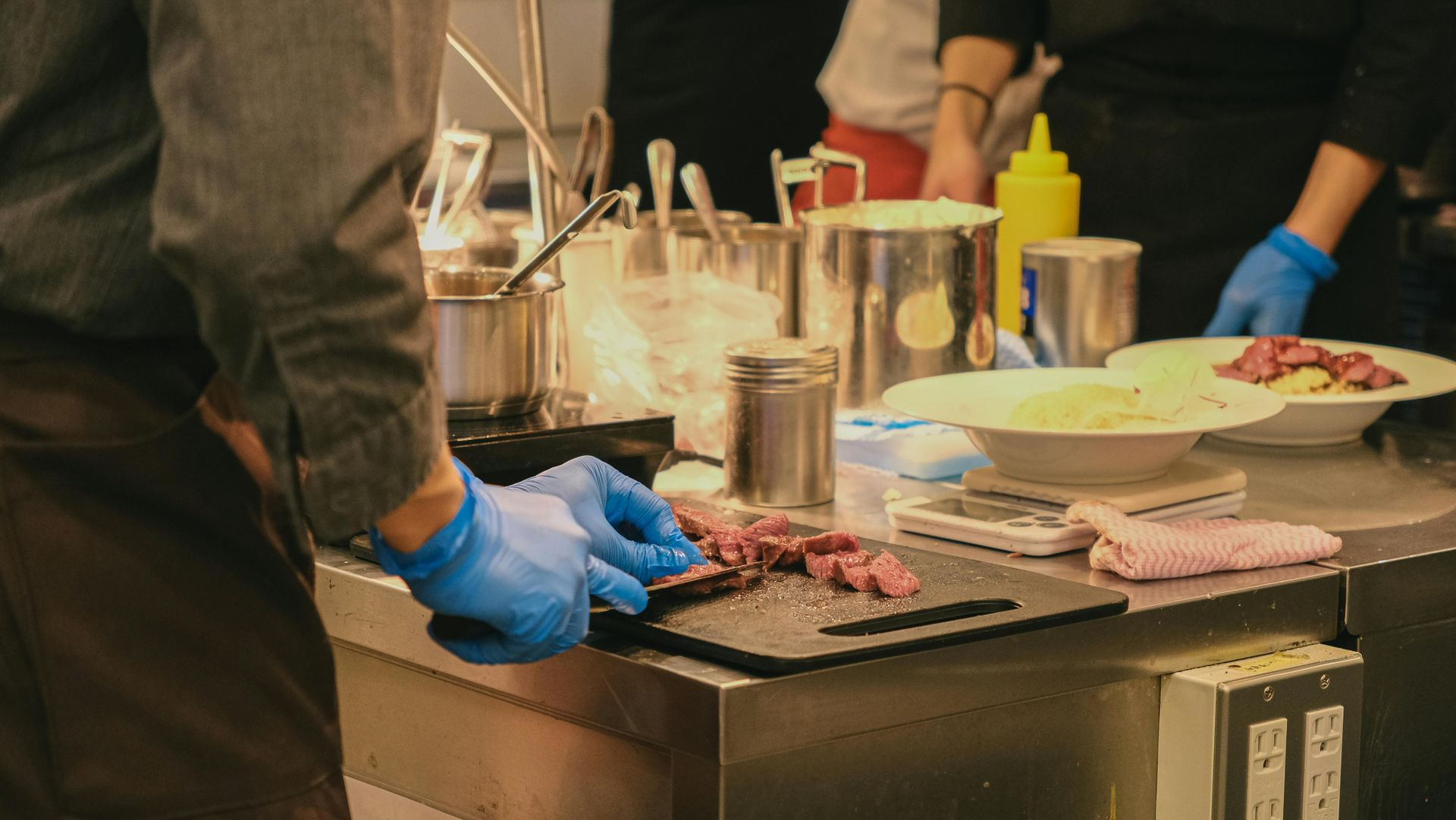 A chef in a professional kitchen uses blue gloves to slice meat on a cutting board near prepared plates.