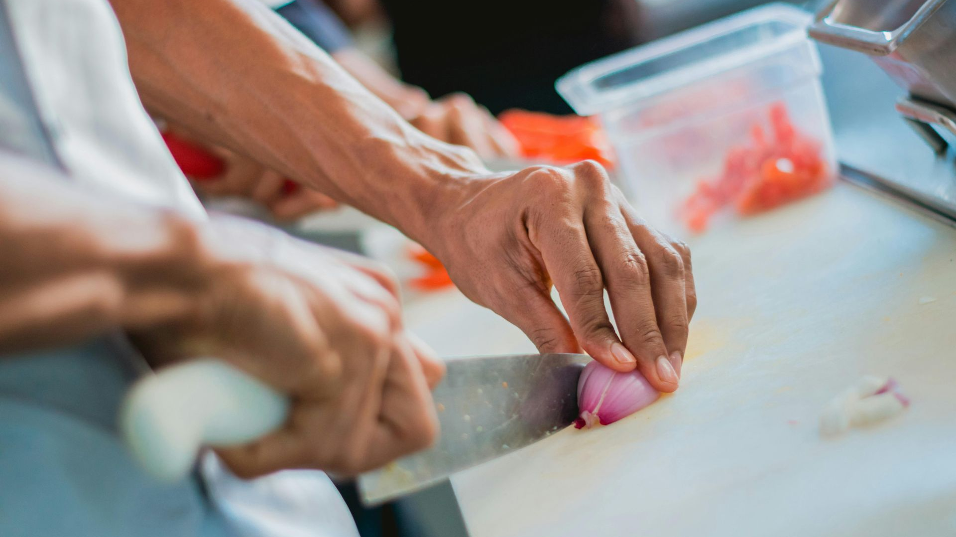 A chef's hands use a large knife to slice a red onion on a white cutting board in a professional kitchen.