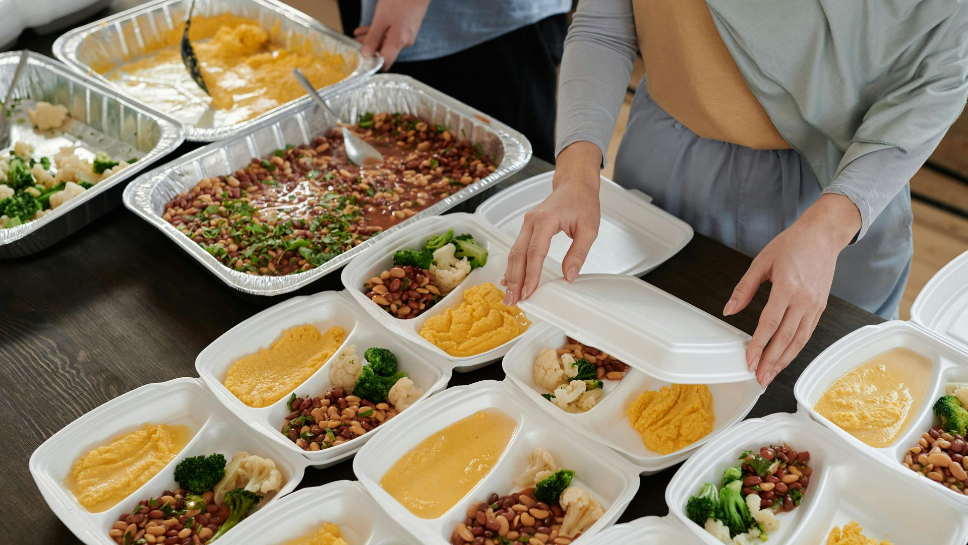 People packing meals into disposable containers from large trays of food at a table.