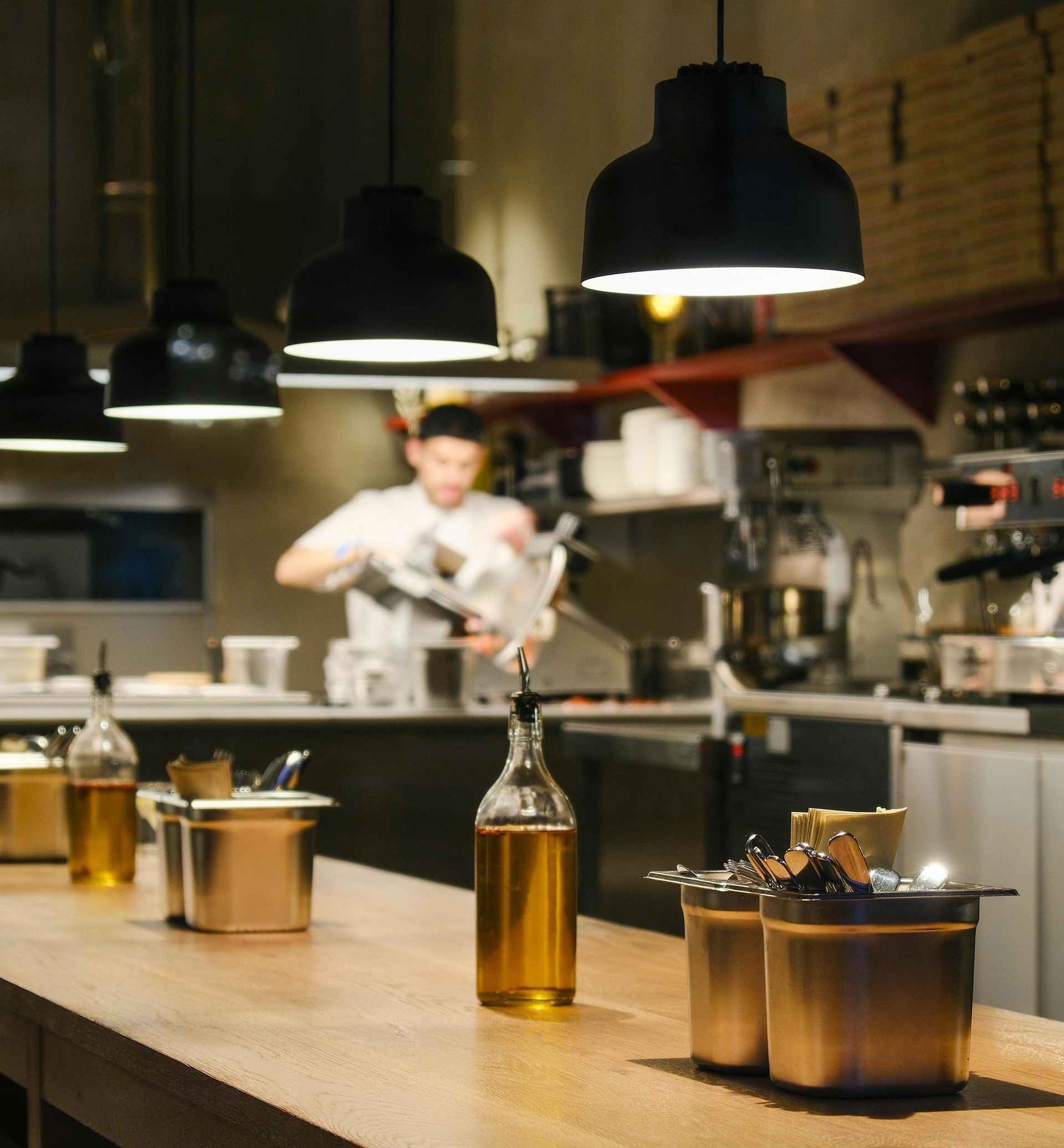 A chef works in a kitchen behind a wooden table topped with oil bottles and metal containers under hanging lights.