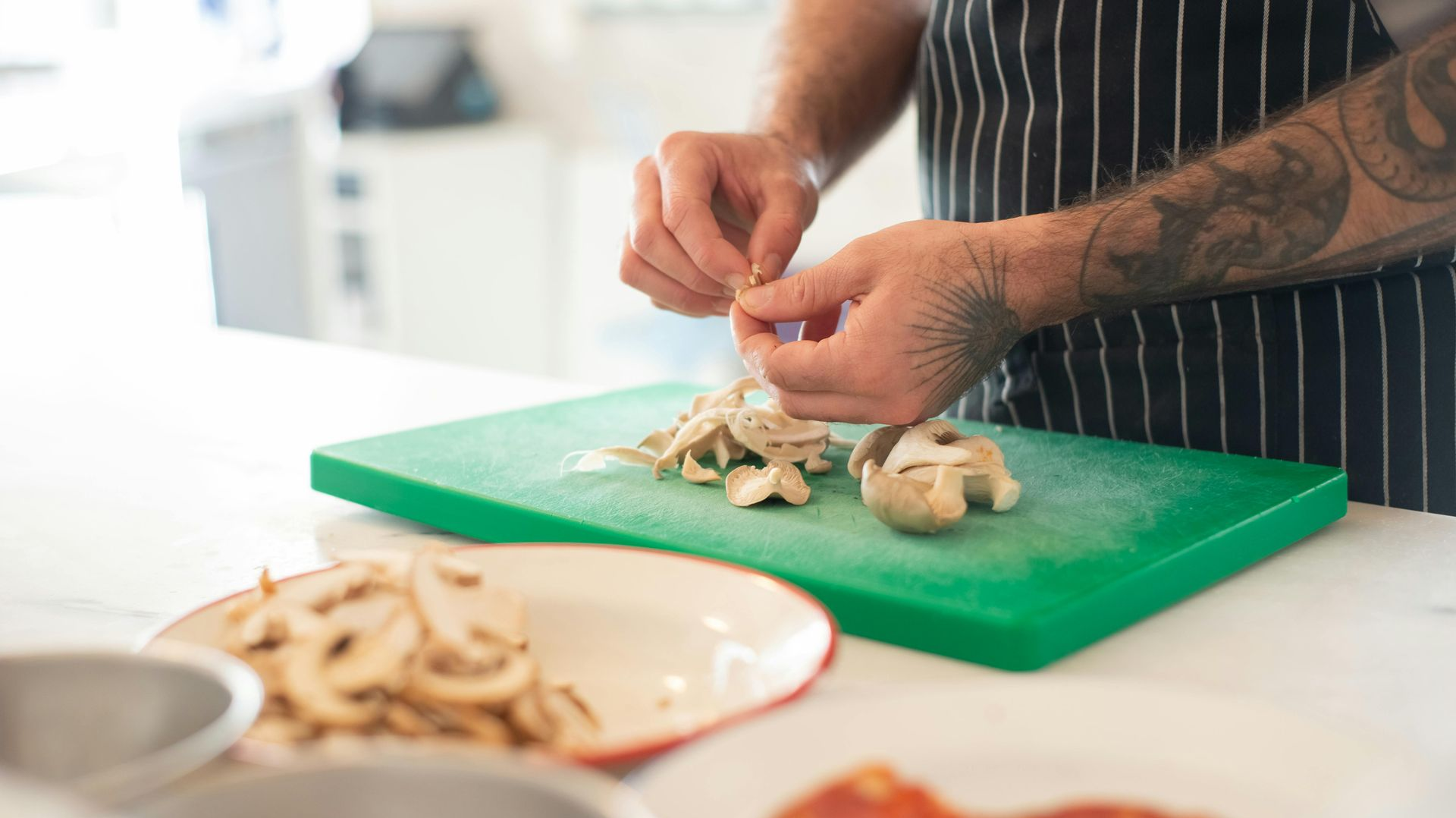 A person with tattoos on their arm prepares sliced mushrooms on a green cutting board in a kitchen.