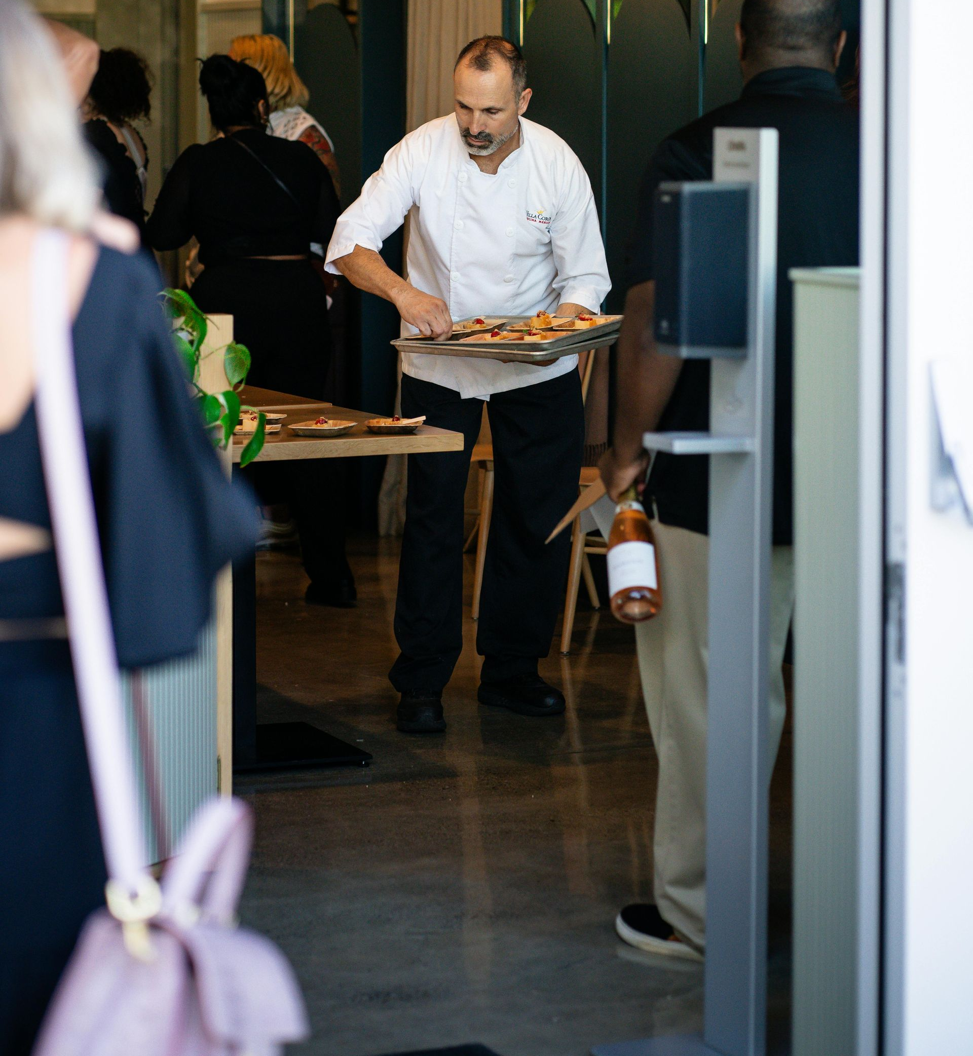 Chef in white uniform carrying tray of food, entering a doorway. People in background.
