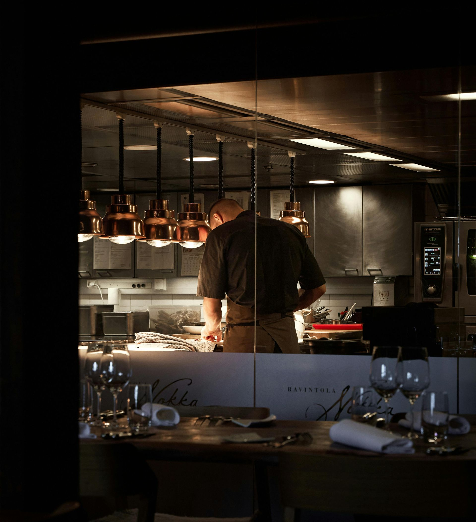 Chef working in a restaurant kitchen, viewed through a window. Copper-toned lights hang above a stainless steel counter.