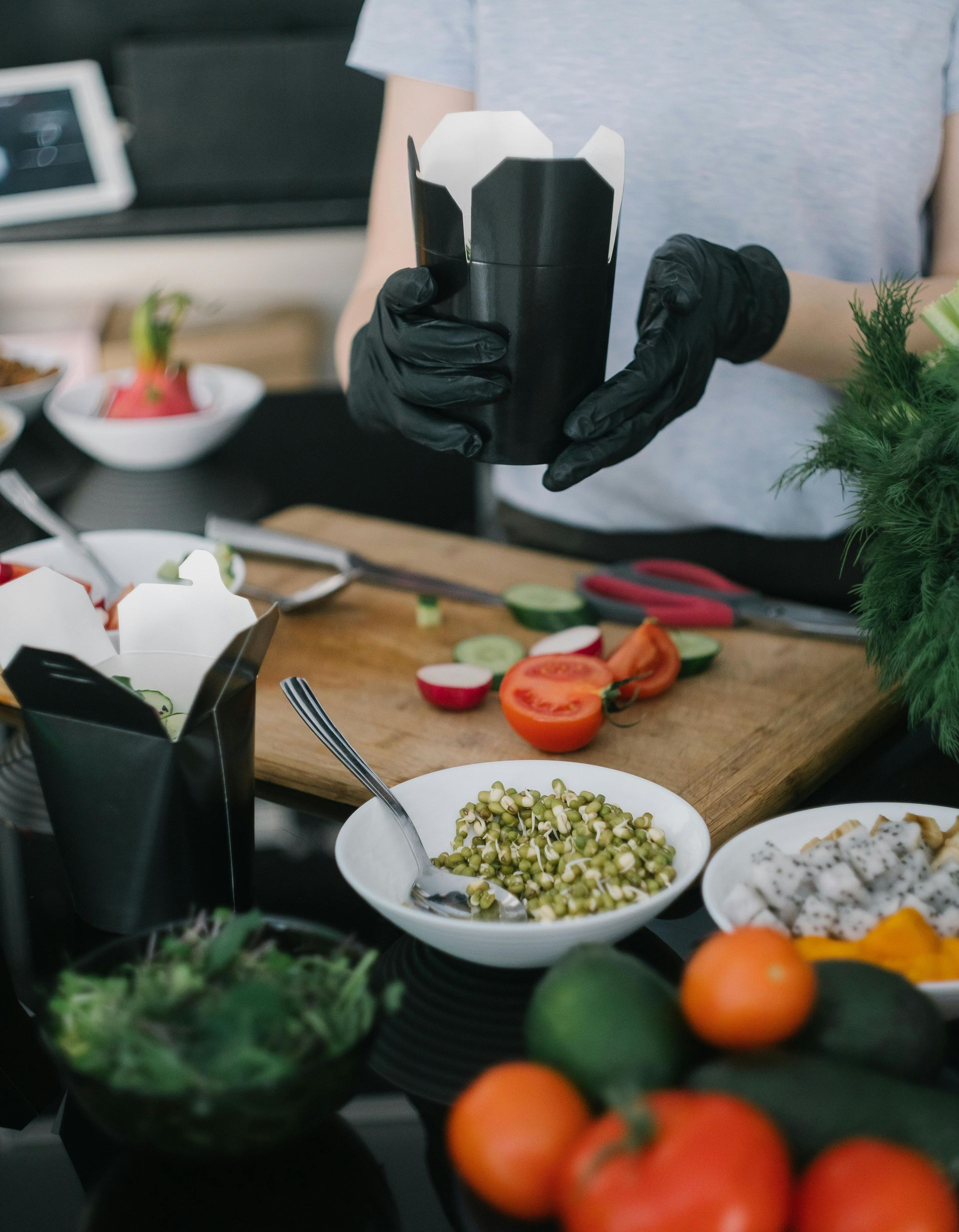A person wearing black gloves holds a black takeout container above a wooden cutting board filled with sliced vegetables.