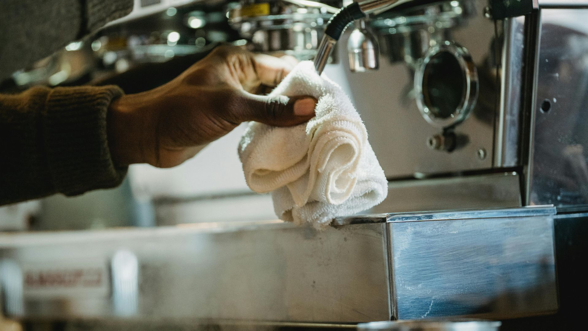 Person wiping a steaming coffee machine nozzle with a white cloth.