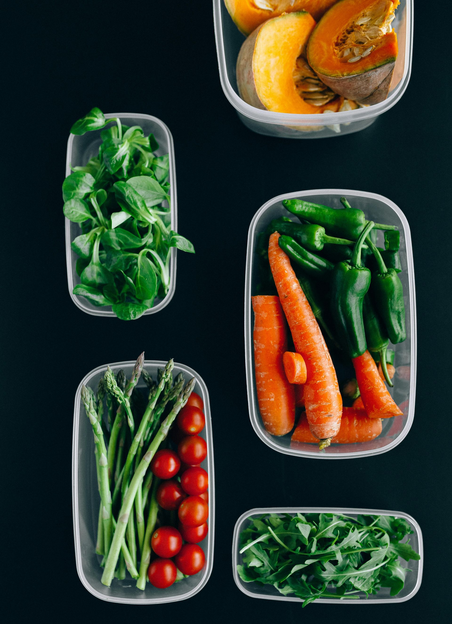 Five clear plastic containers filled with fresh vegetables, including squash, greens, carrots, peppers, and tomatoes.