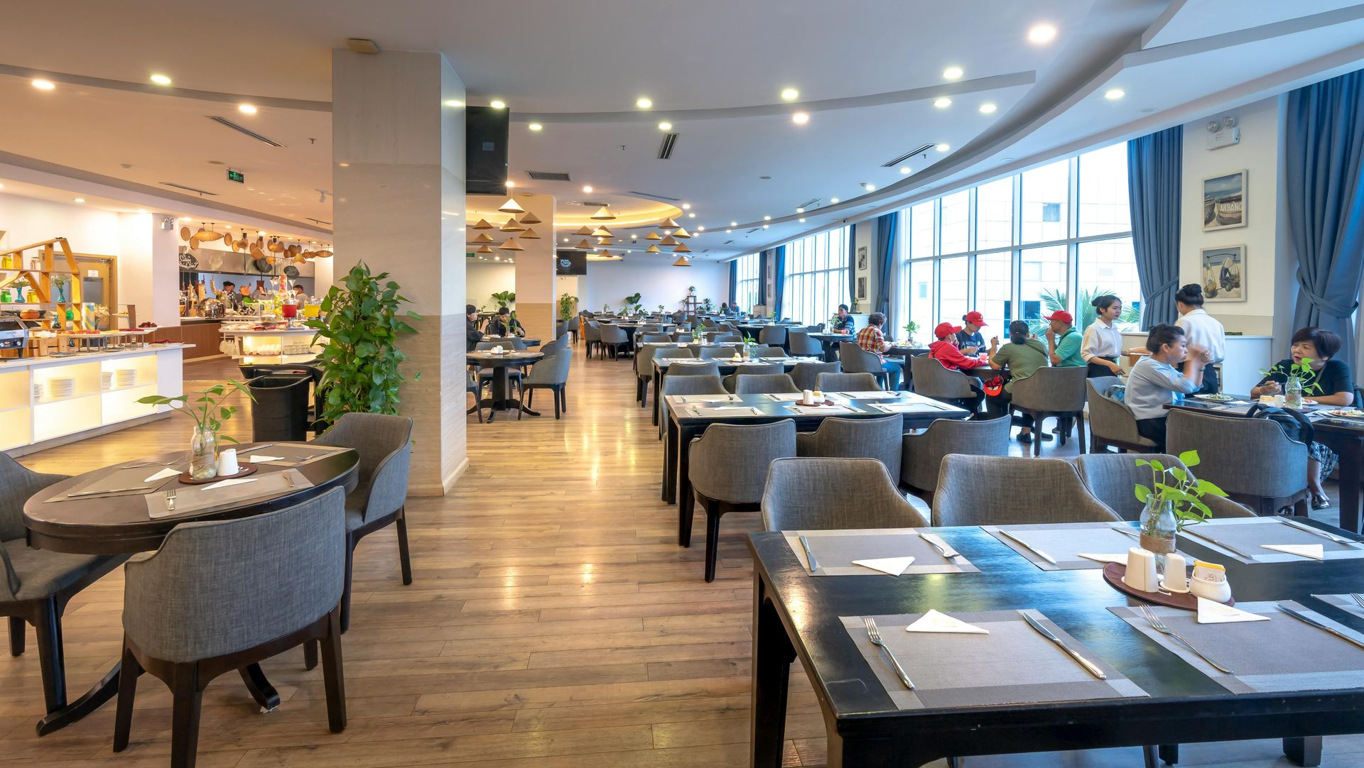 Restaurant interior with tables, chairs, and buffet area. Natural light streams in through large windows.