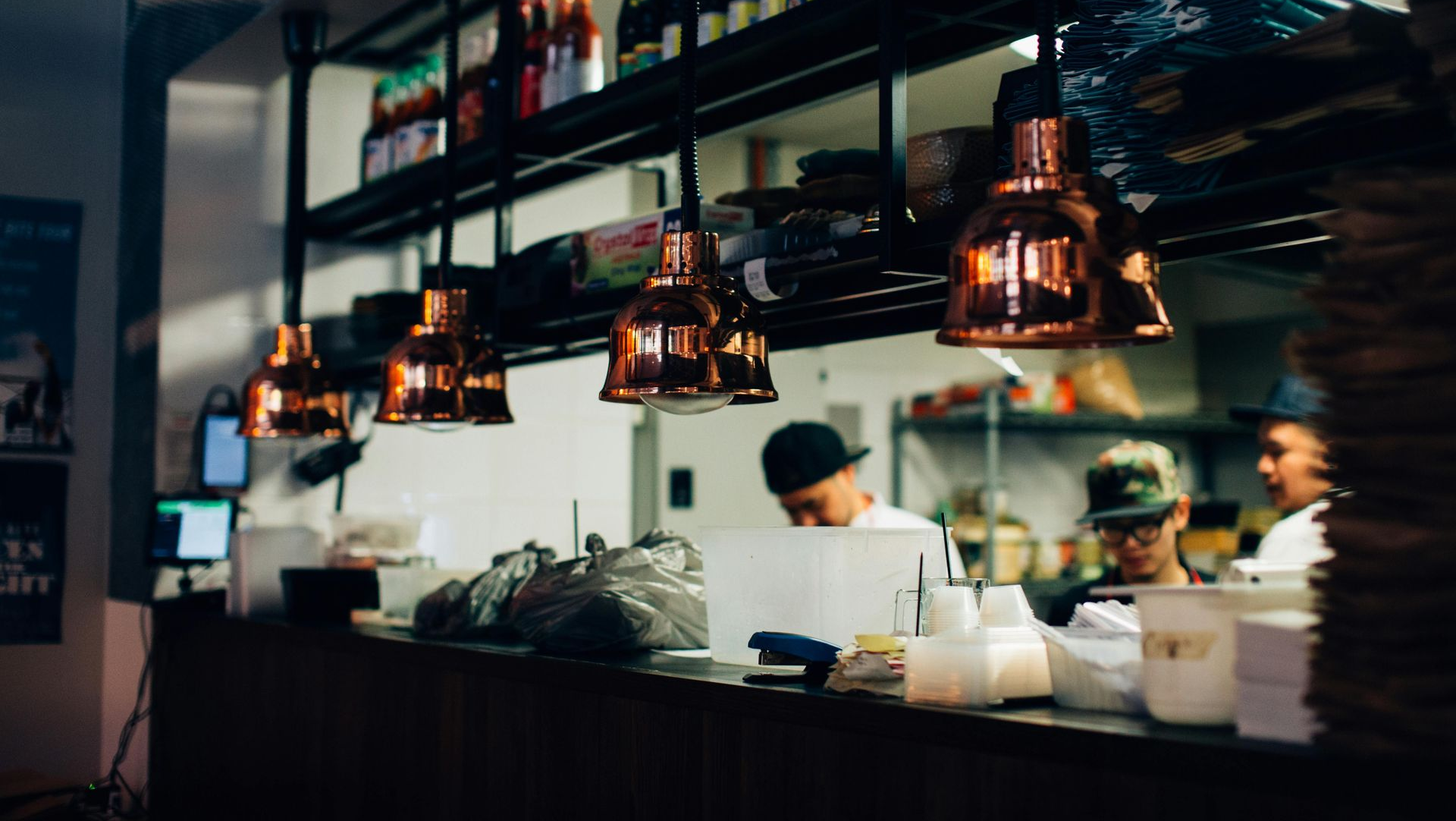 Copper pendant lights illuminate a restaurant's kitchen counter where staff work.