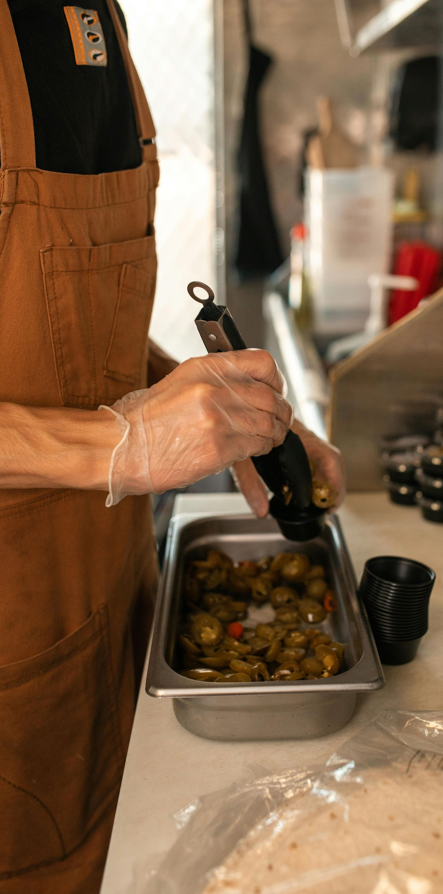 Person in apron using tongs to put jalapeños from a container into a tortilla.