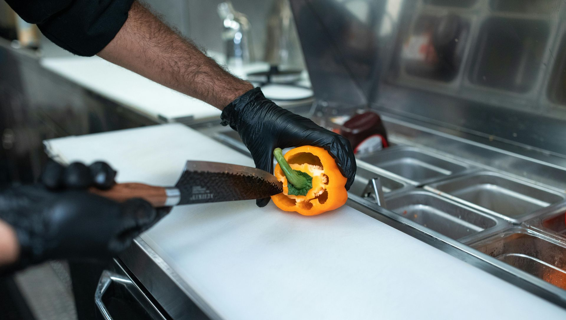 Person wearing black gloves slicing a yellow bell pepper on a white cutting board near a food prep station.