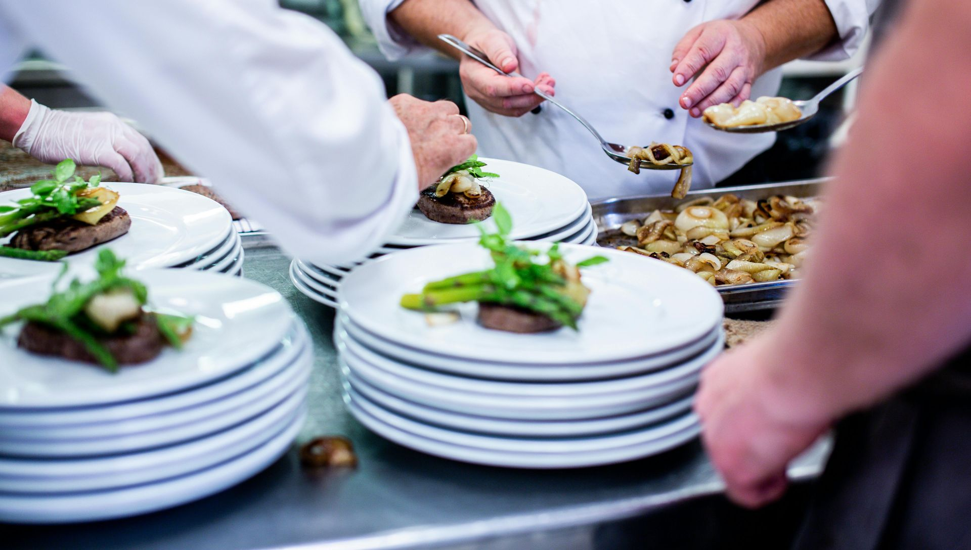 Chefs plating gourmet meals in a commercial kitchen; white plates, food, stainless steel.