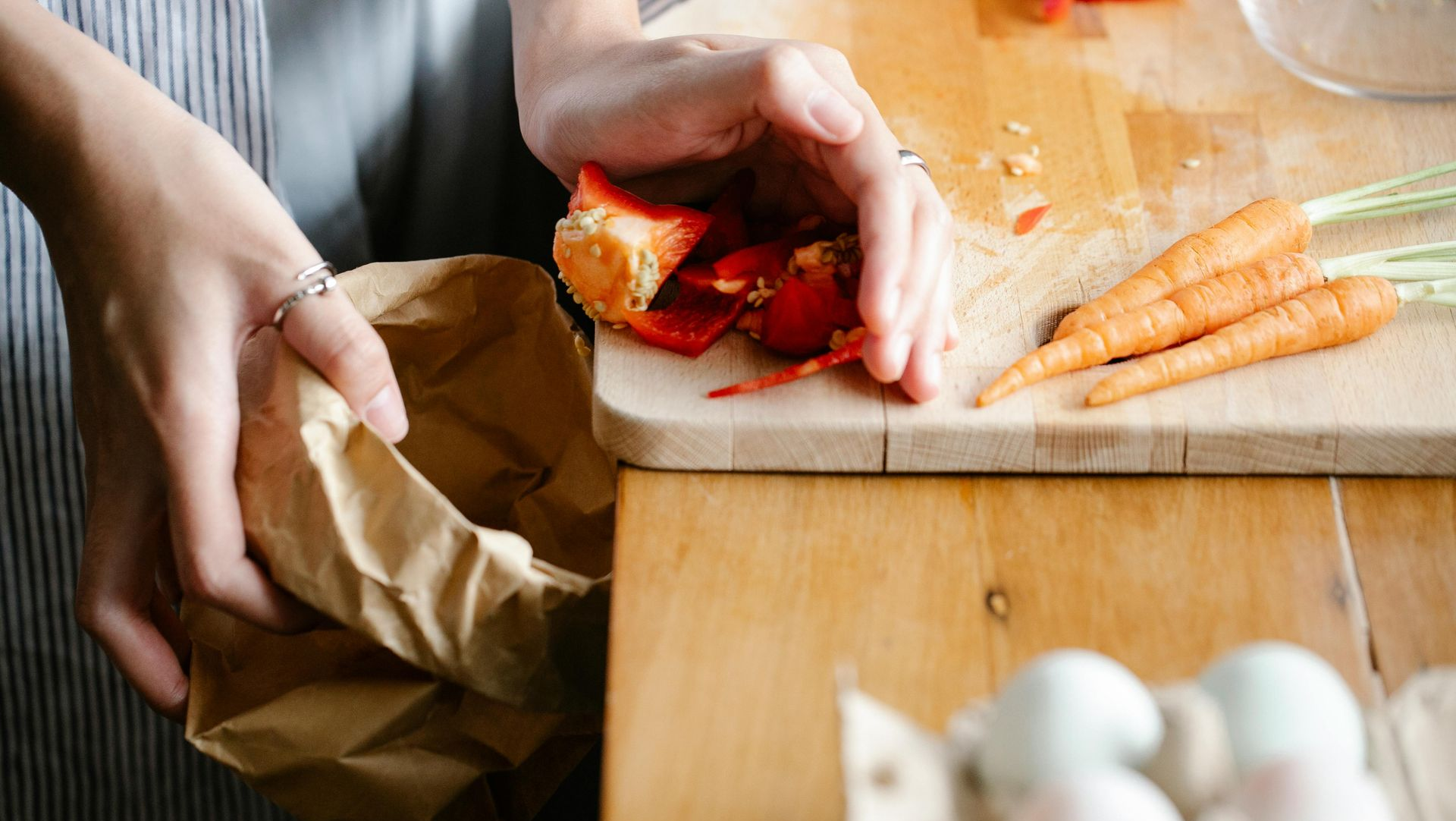 A person holding a brown paper bag while preparing red bell peppers and small carrots on a wooden kitchen cutting board.