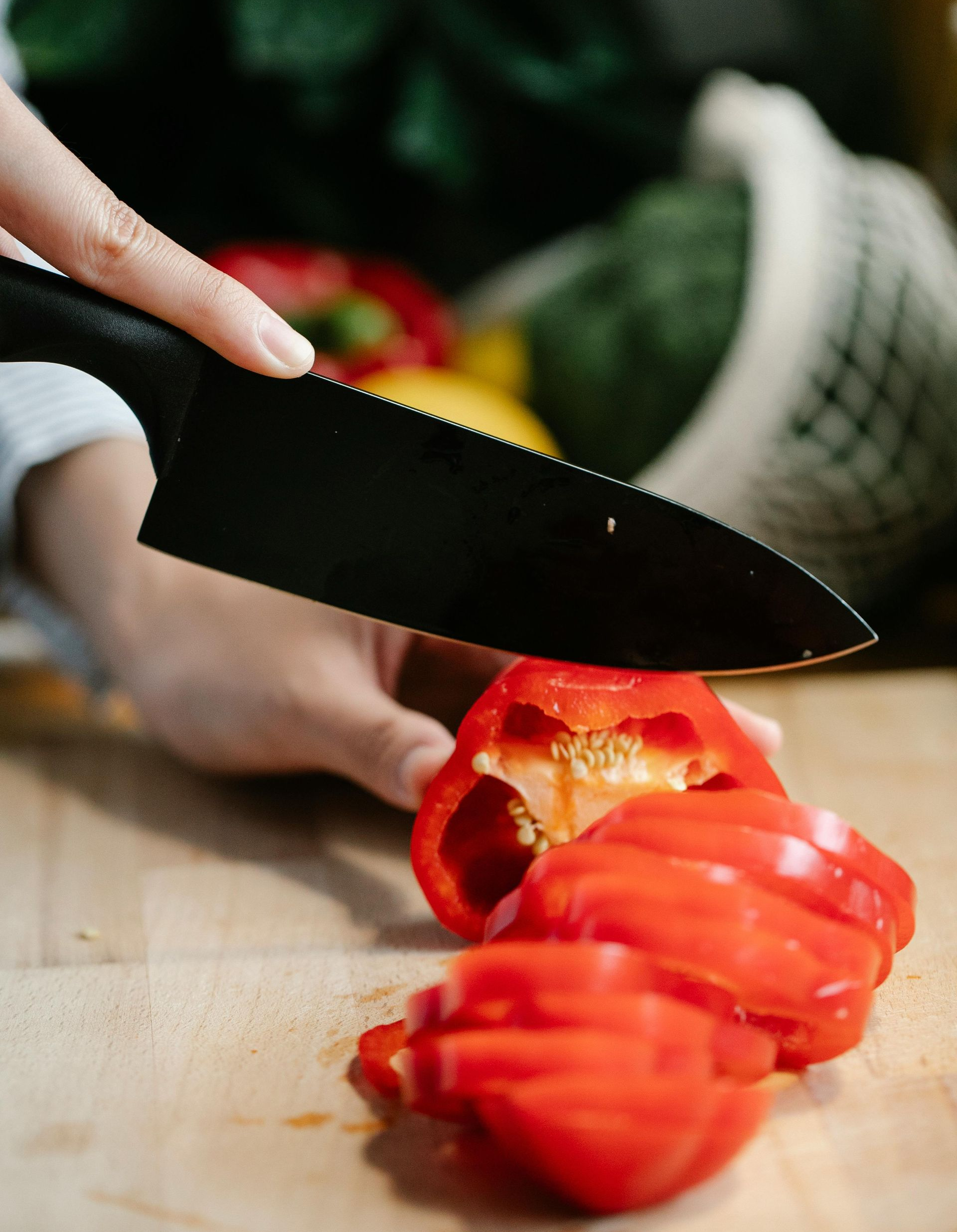 A person uses a black serrated knife to slice a red bell pepper on a wooden cutting board.