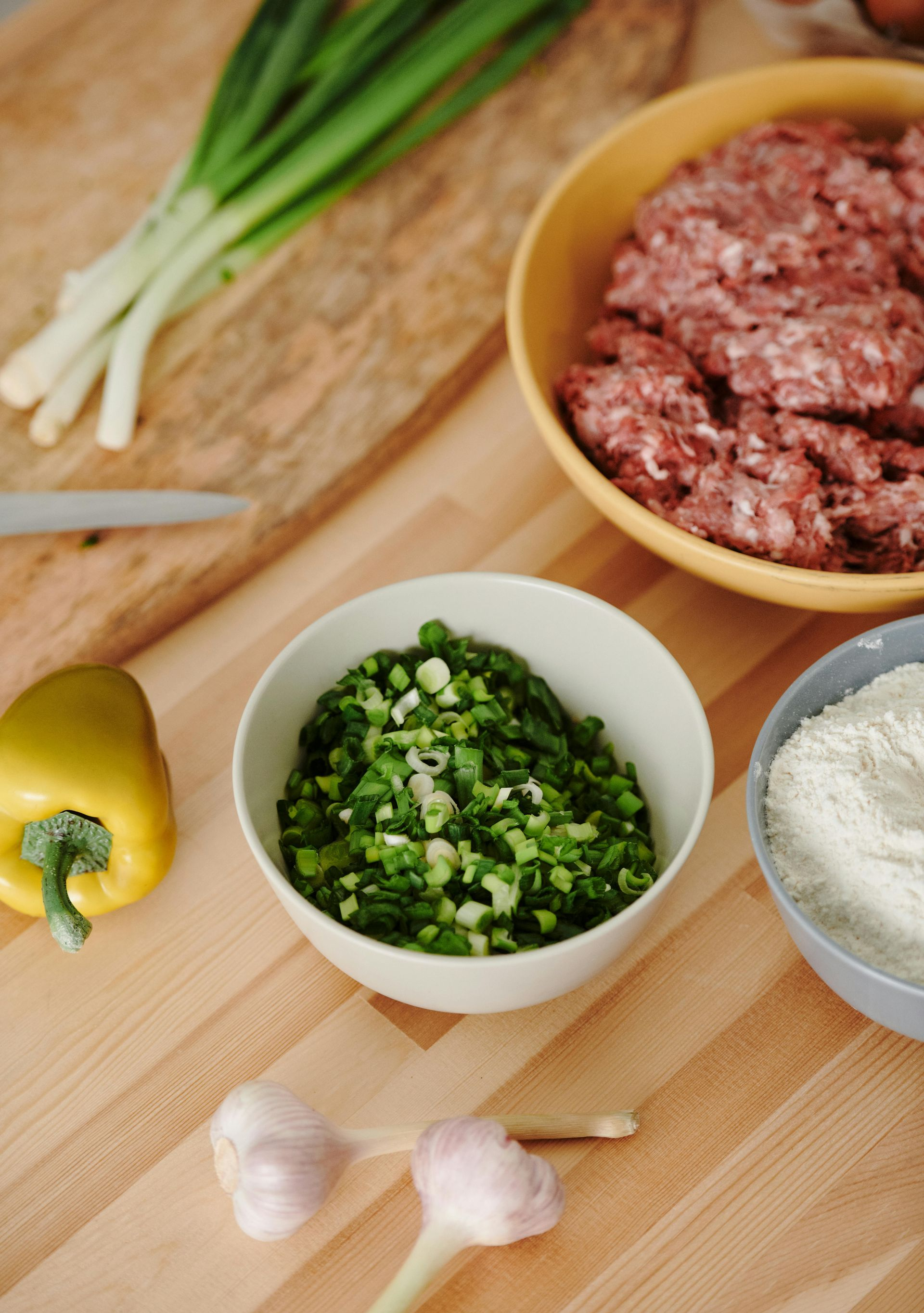 Ingredients for cooking including minced meat, flour, chopped green onions, a yellow pepper, and garlic on a wooden table.