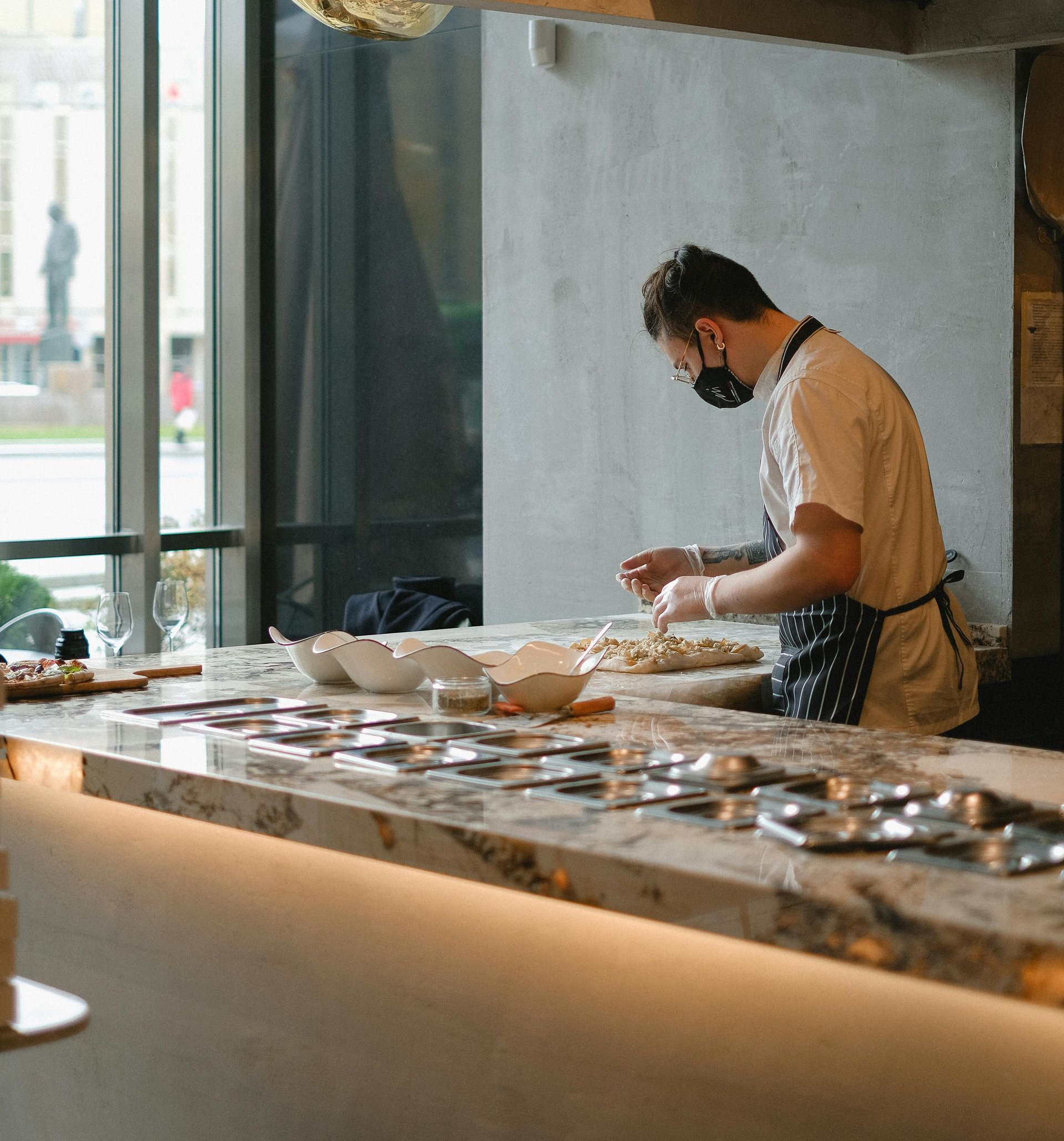 Chef in apron and mask preparing food at a marble countertop in a well-lit kitchen.