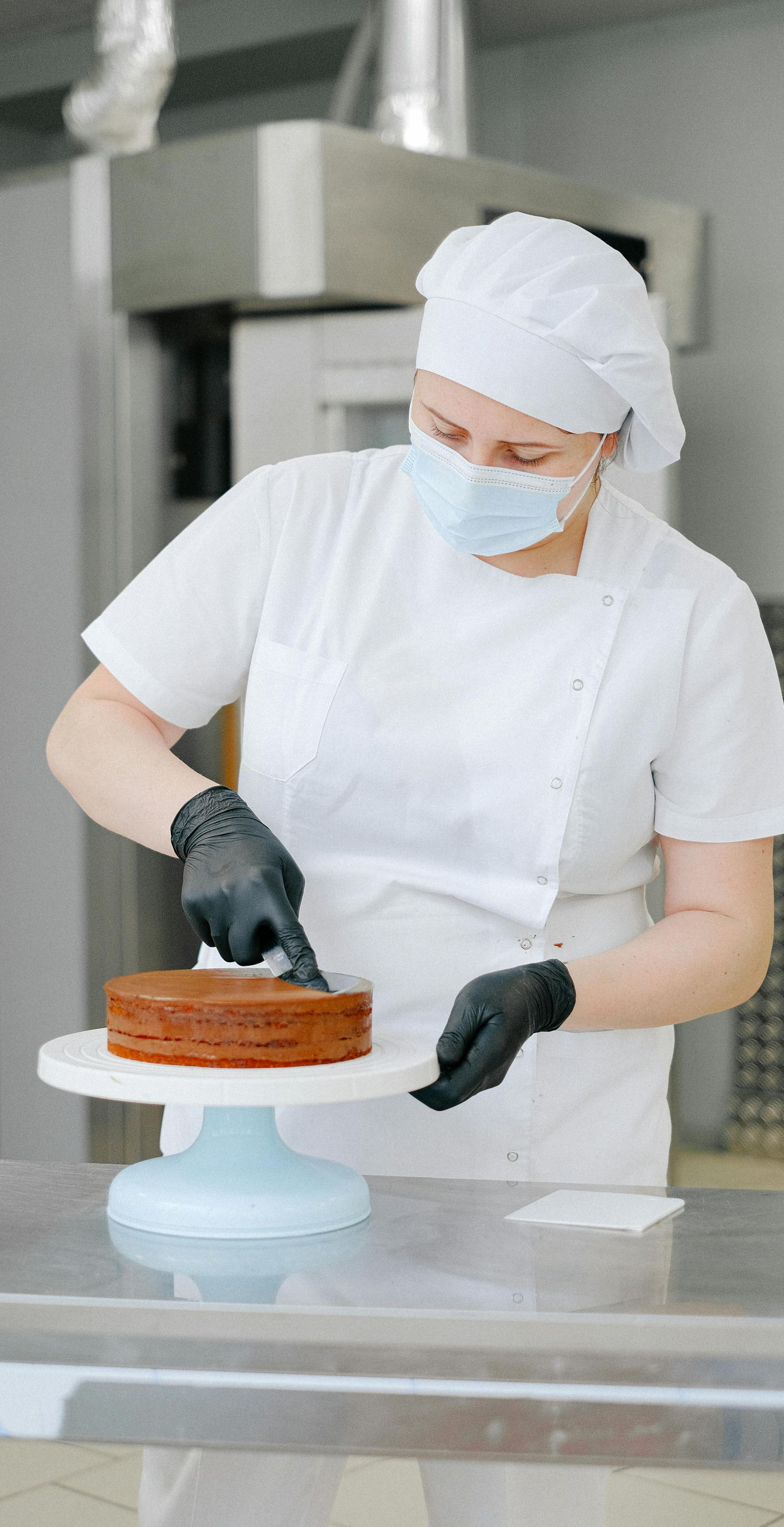 Baker in a white uniform and face mask, preparing a cake on a stand.