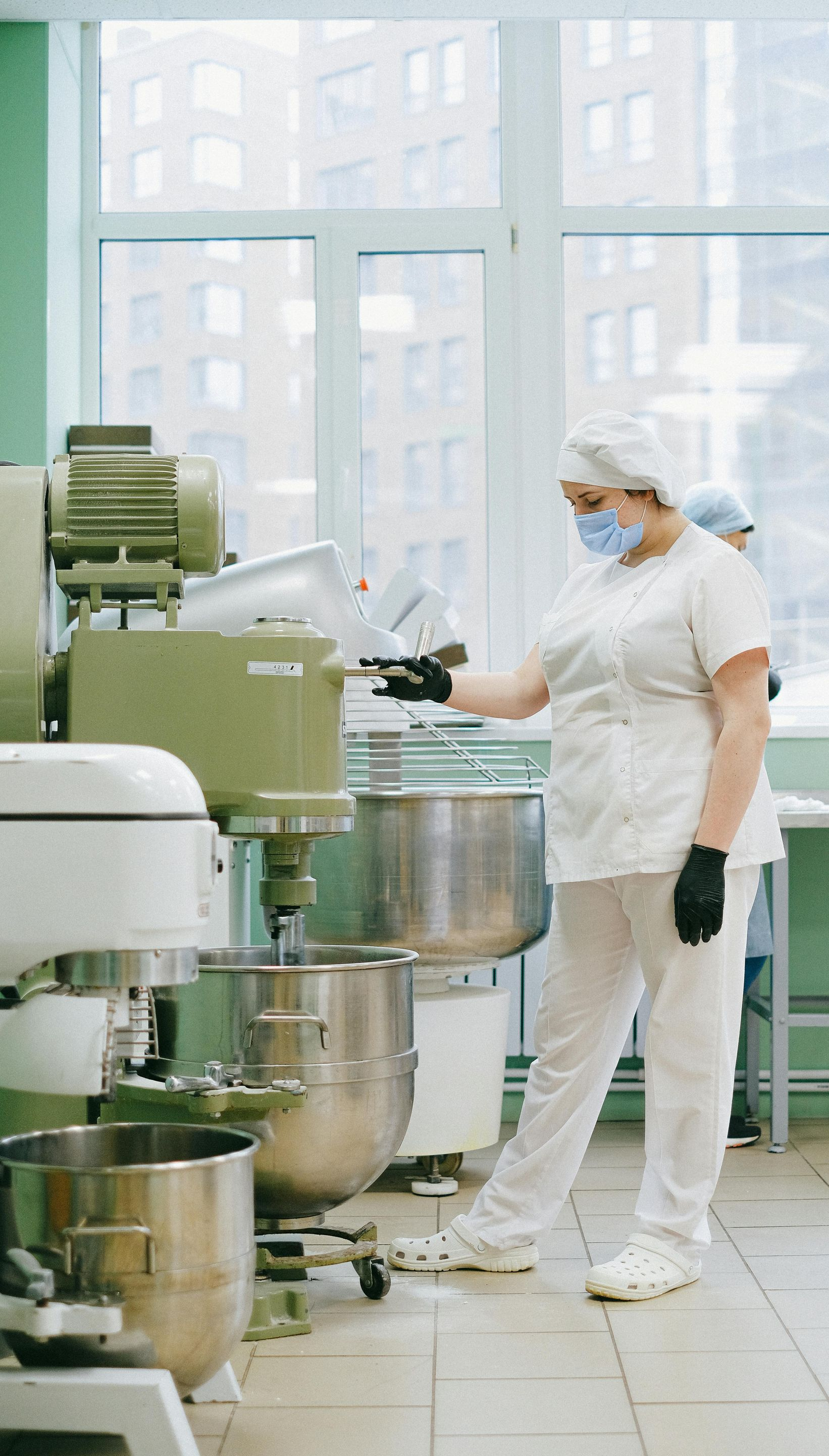 Person in white uniform operates industrial mixer in a bright kitchen.