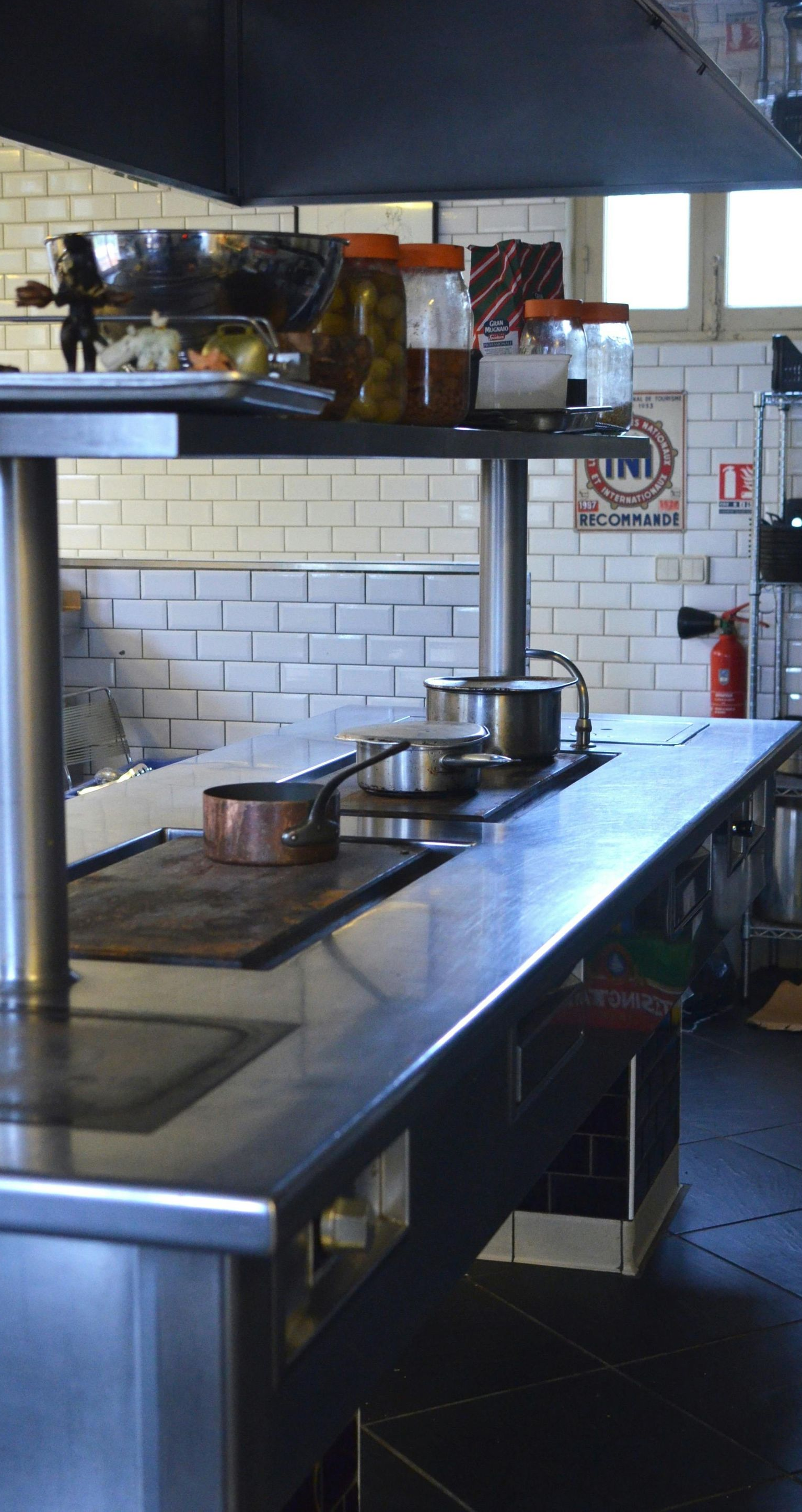 Kitchen with stainless steel stove, pots, and jars on a counter, white tiled walls.