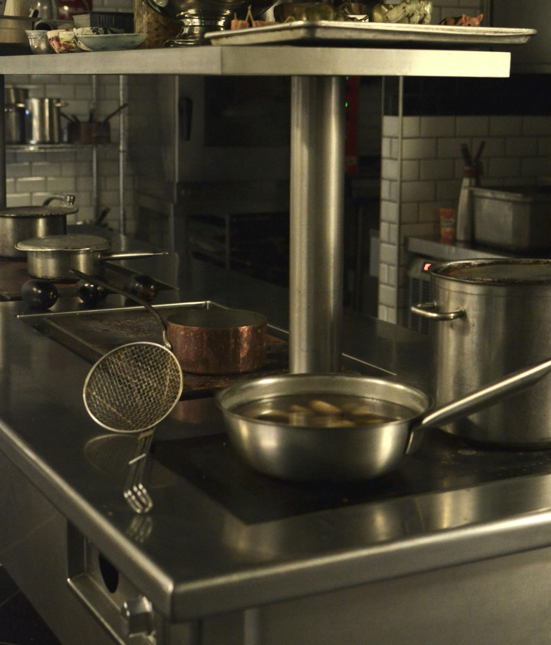 Stainless steel kitchen counter with various pots, pans, and a skimmer utensil, set in a professional restaurant kitchen.