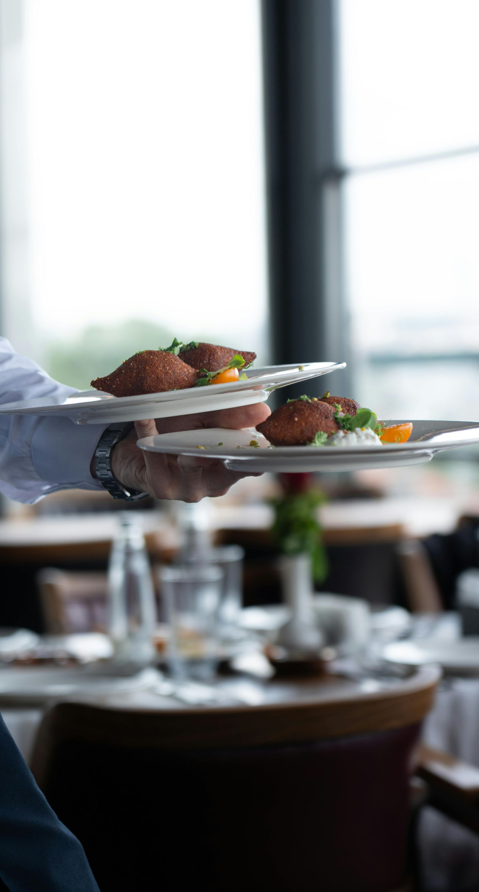 Server carrying two plates of food in a restaurant, near a window.