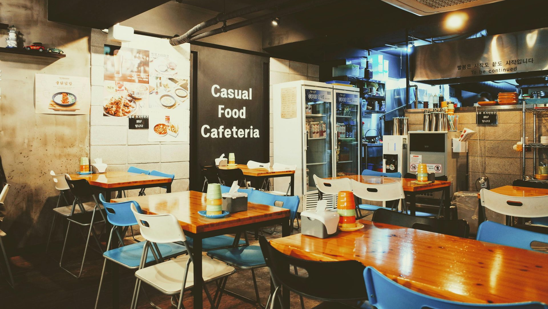 A small cafeteria interior with wooden tables and colorful chairs under warm lighting, featuring a