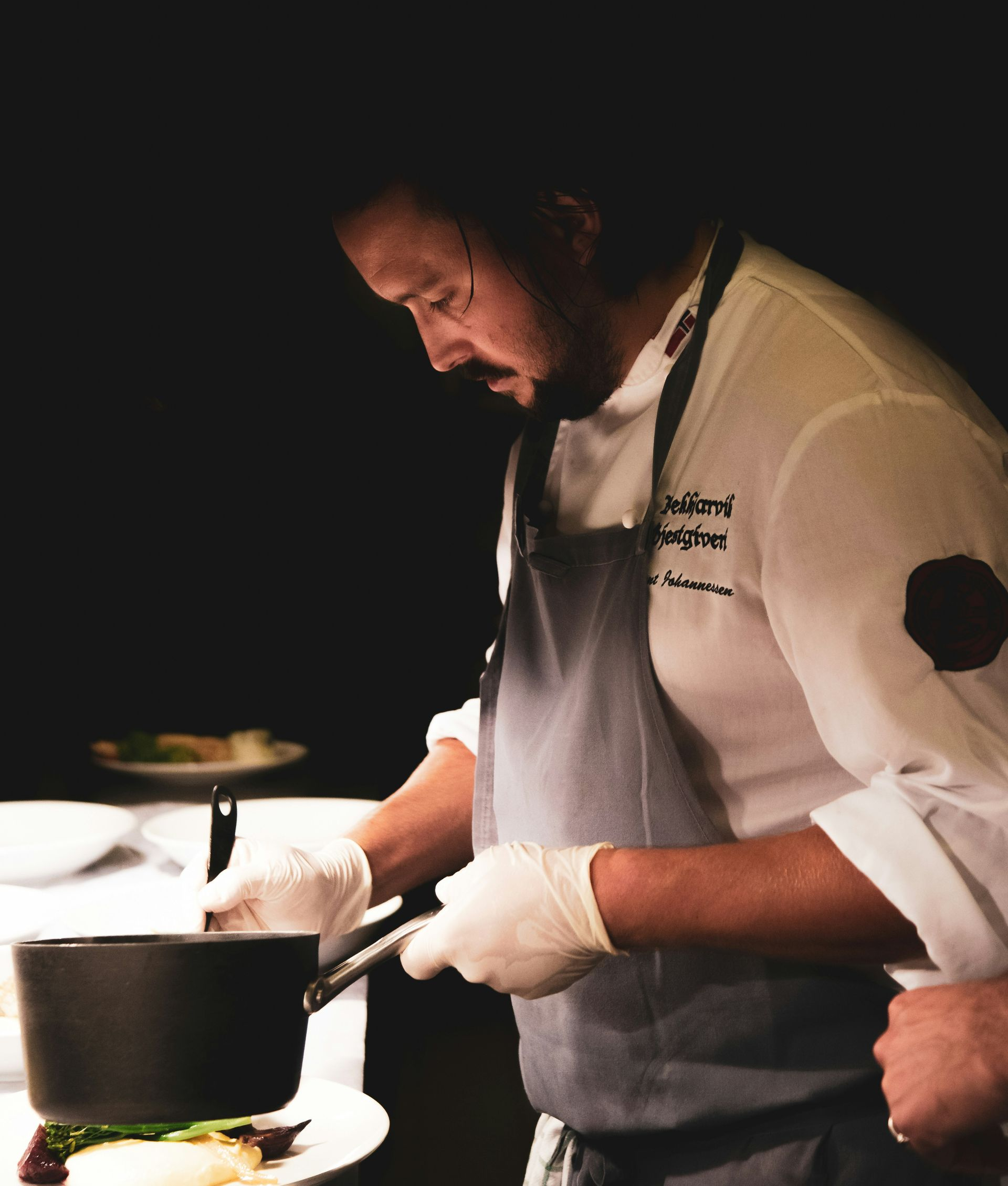 Chef wearing apron and gloves leaning over a pot on a stovetop, focused and working.