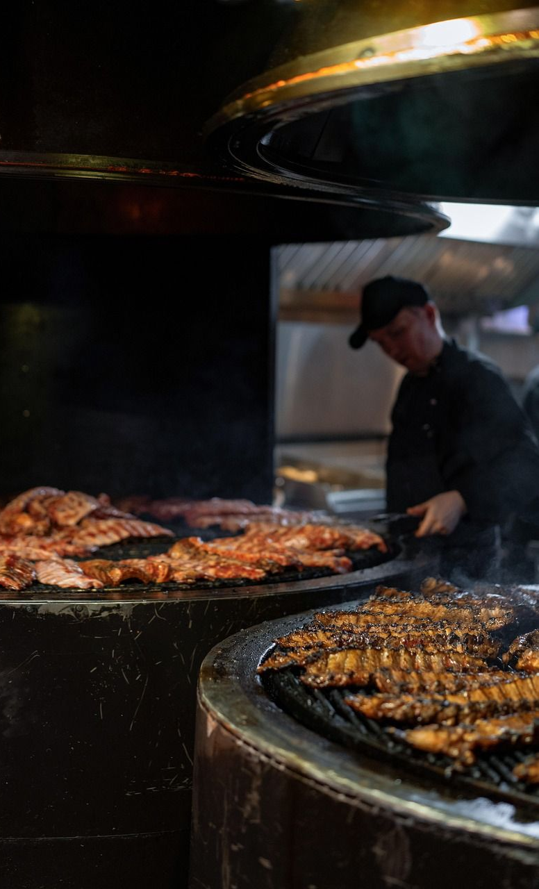 Chef grilling racks of ribs in a commercial kitchen with large smoker.
