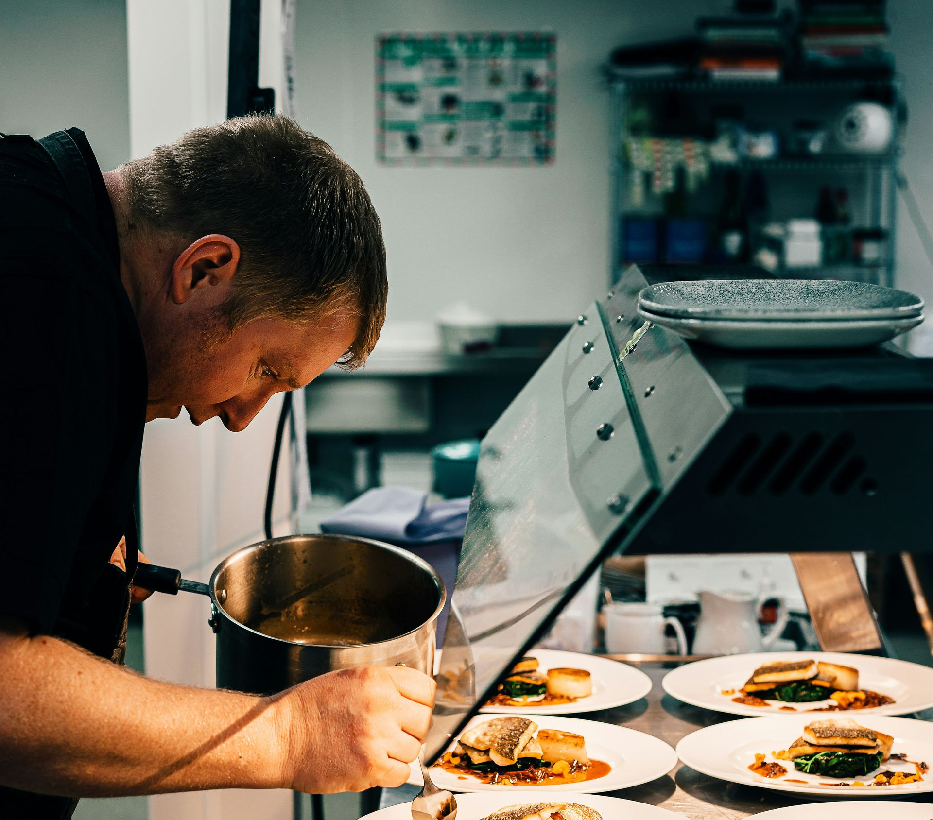 Chef pours sauce on plated dishes in a commercial kitchen.