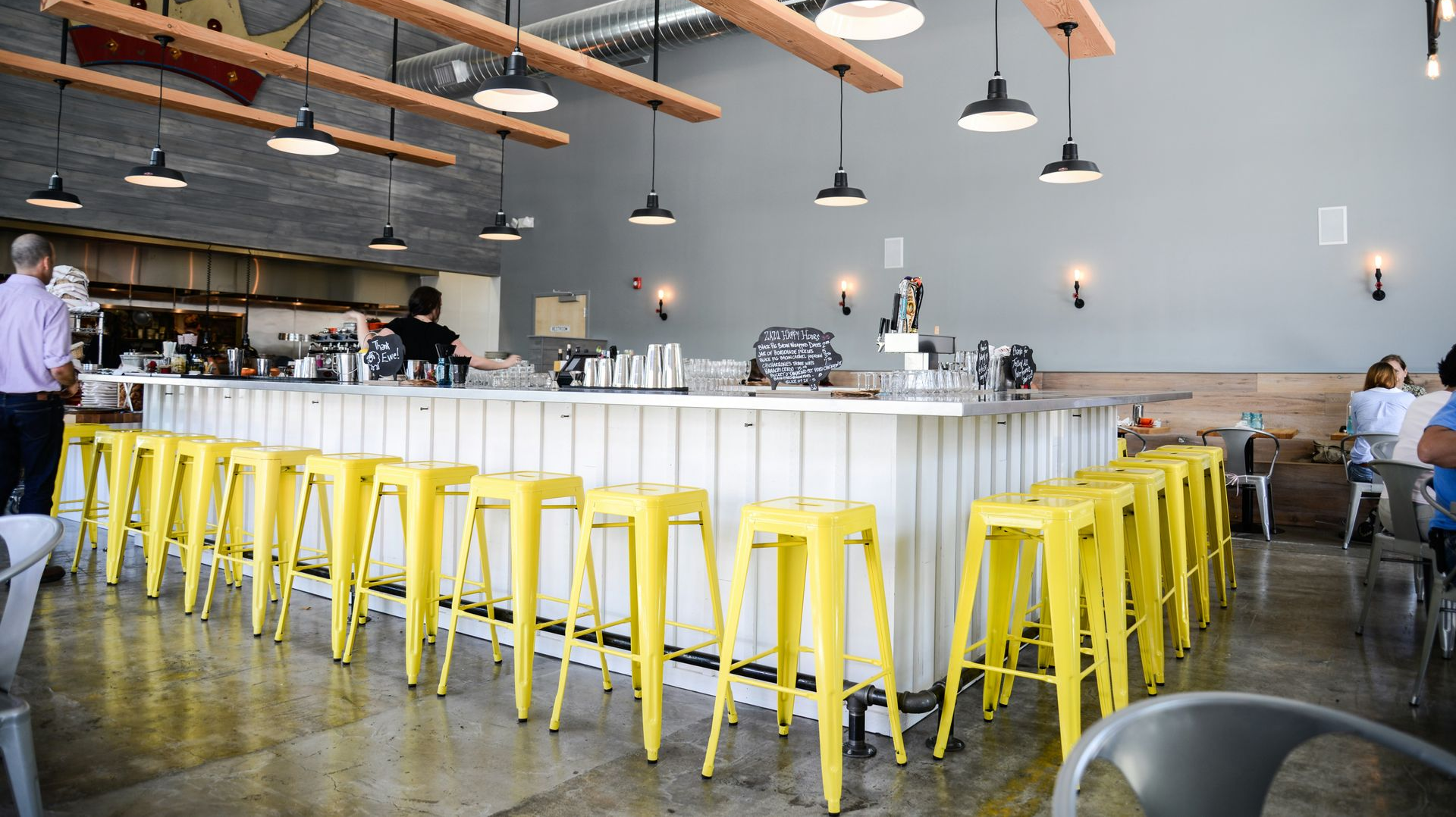 Modern cafe with long white communal table, yellow stools, hanging lights, and exposed wood beams