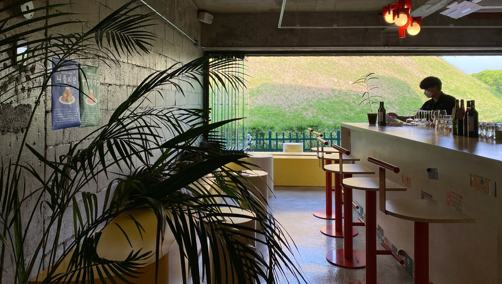 Modern bar interior with a curved counter, hanging lights, and tropical plants overlooking greenery outside