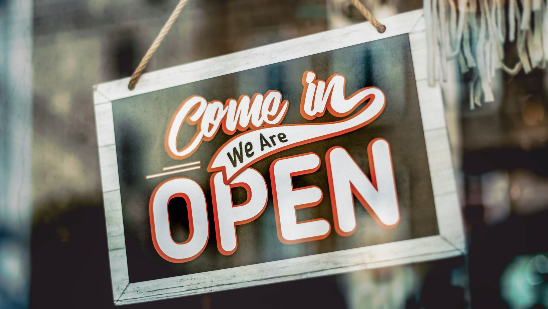 Hanging wooden sign reading “Come in, We Are OPEN” in a storefront window