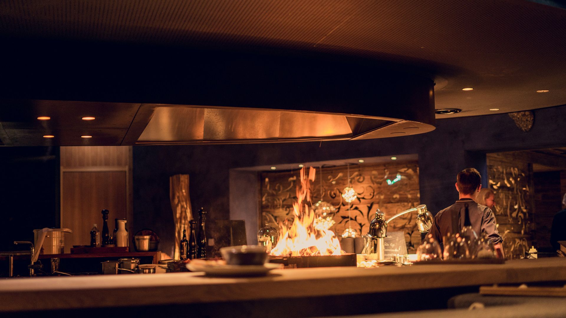 A chef cooks with flames at a restaurant's cooking station, with bottles and plates nearby.