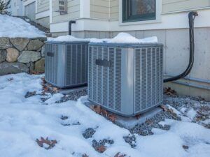 Two air conditioners are sitting in the snow outside of a house.