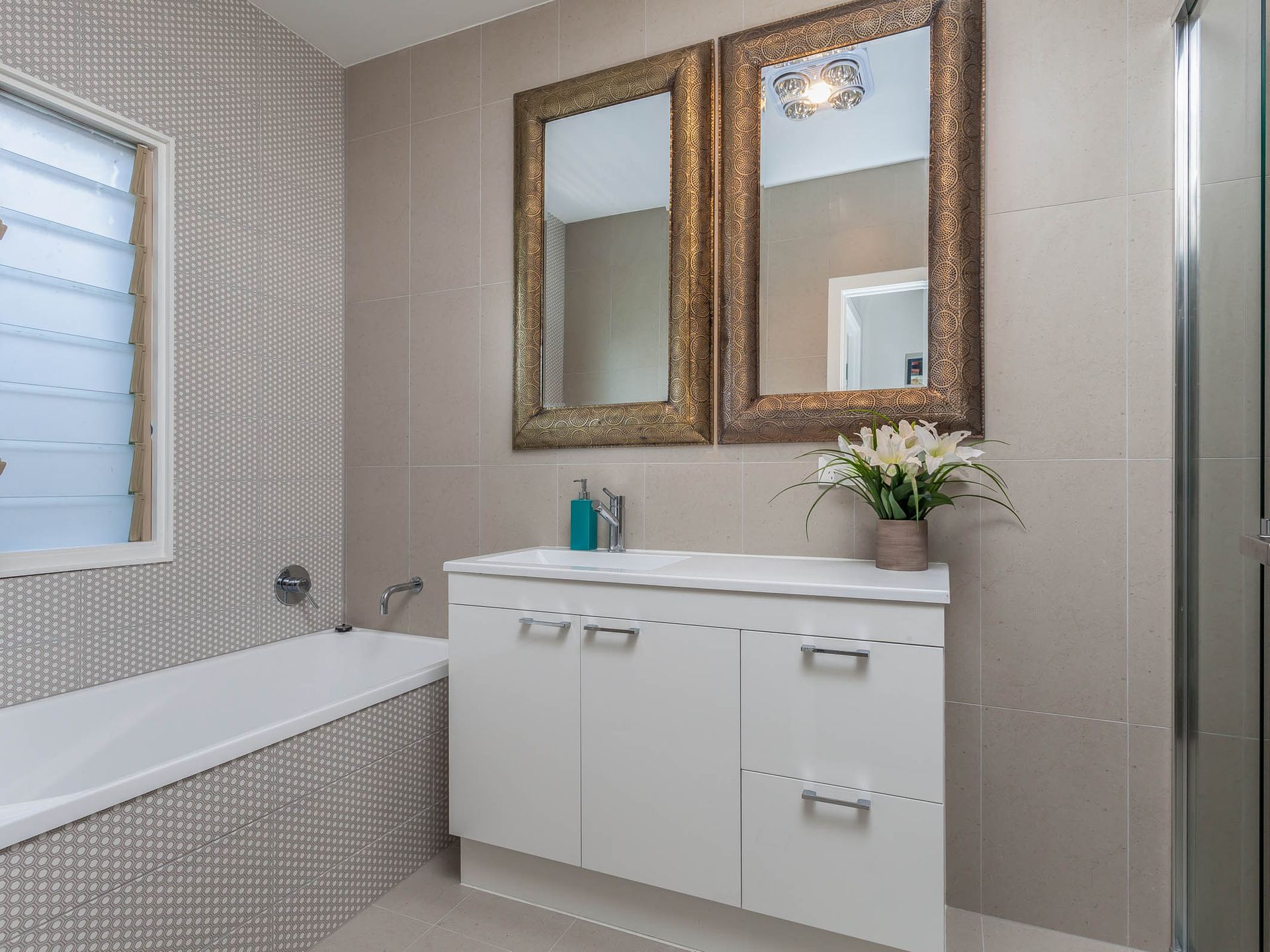 Modern bathroom with a white vanity, gold-framed mirrors, and a white bathtub. The walls are tiled in shades of gray.