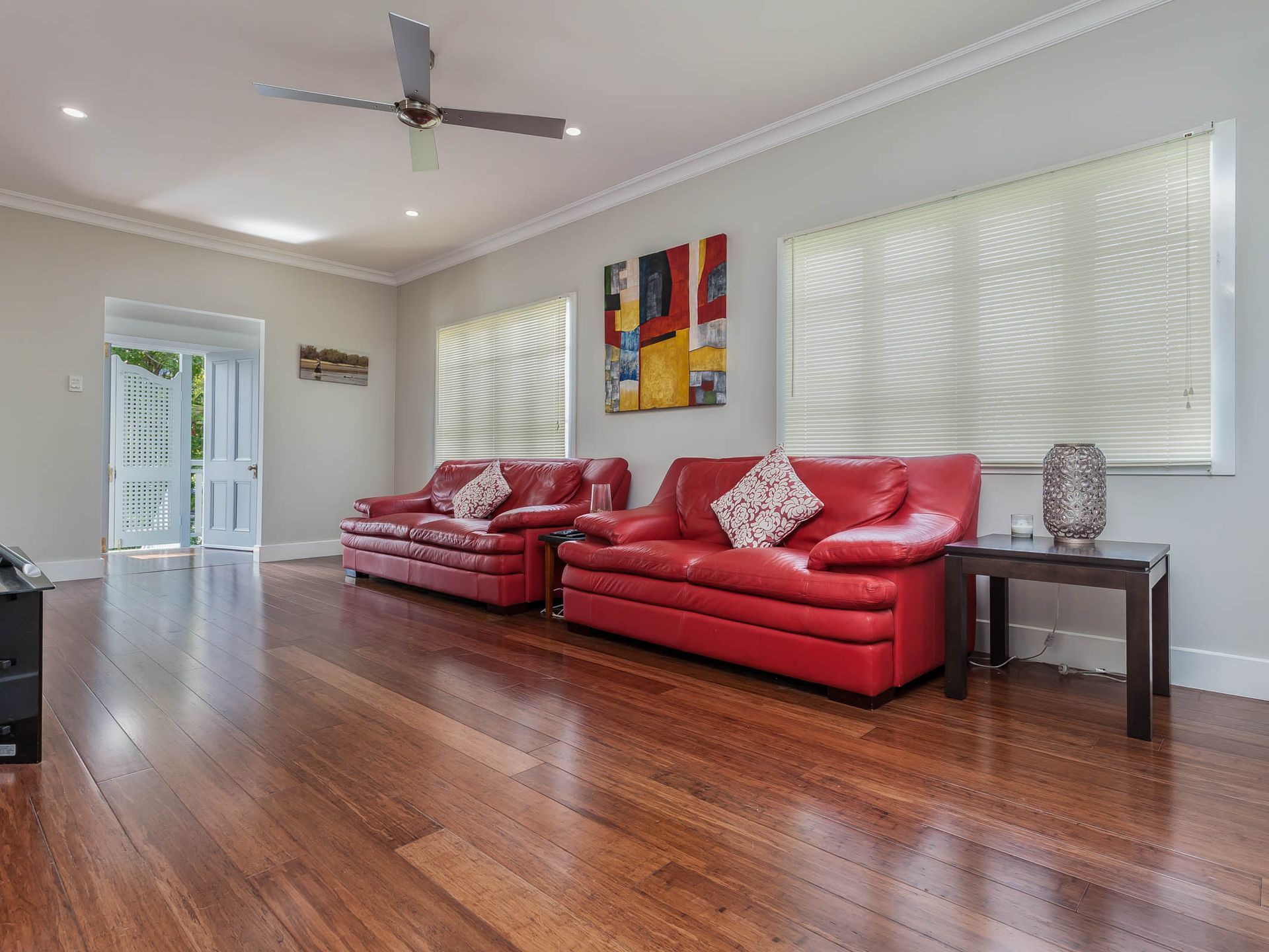 Living room with red leather sofas, hardwood floors, and large windows with blinds. An open door leads outside.
