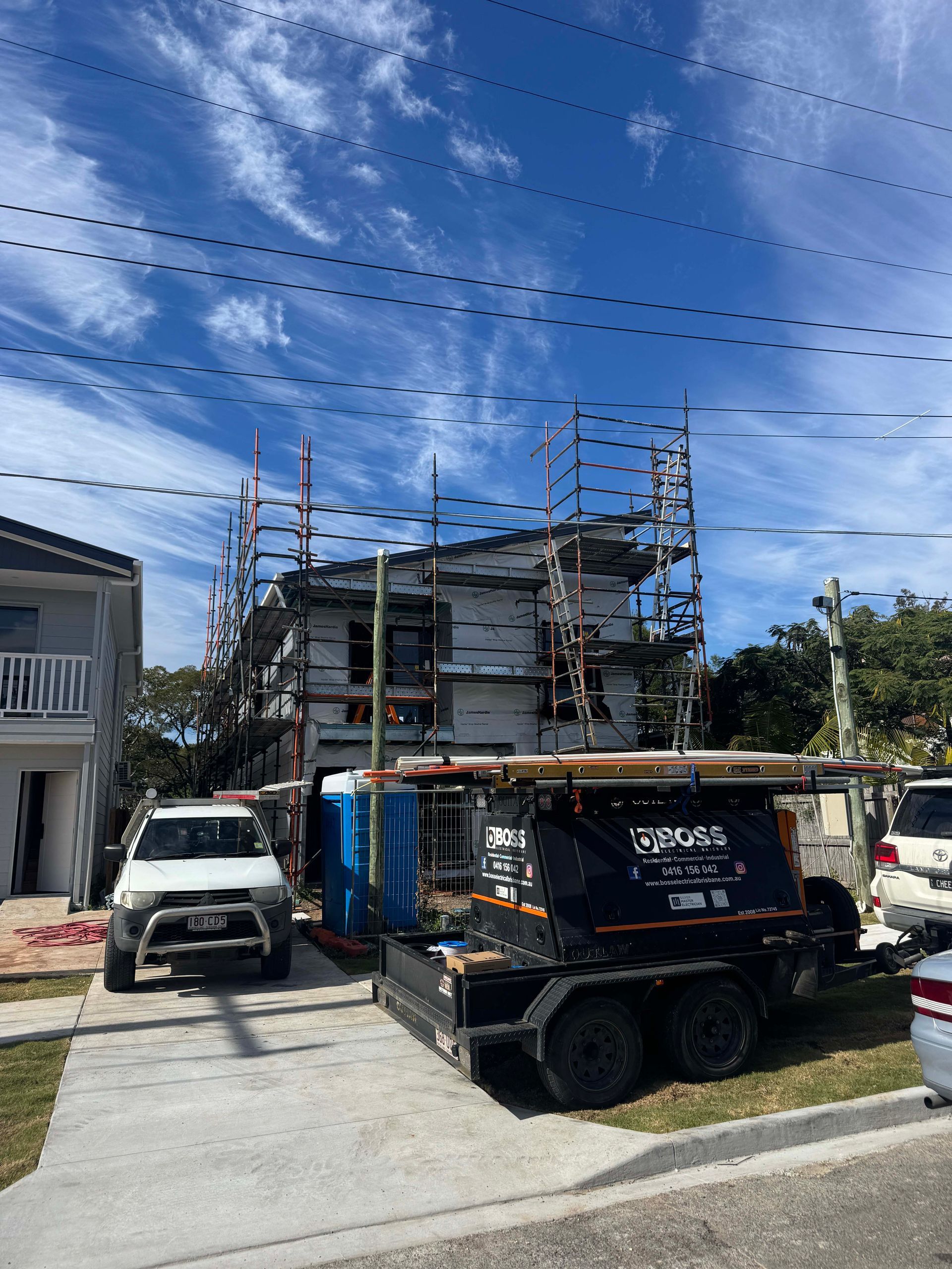 Two-story house under construction with scaffolding, surrounded by vehicles and a portable toilet. Blue sky overhead.