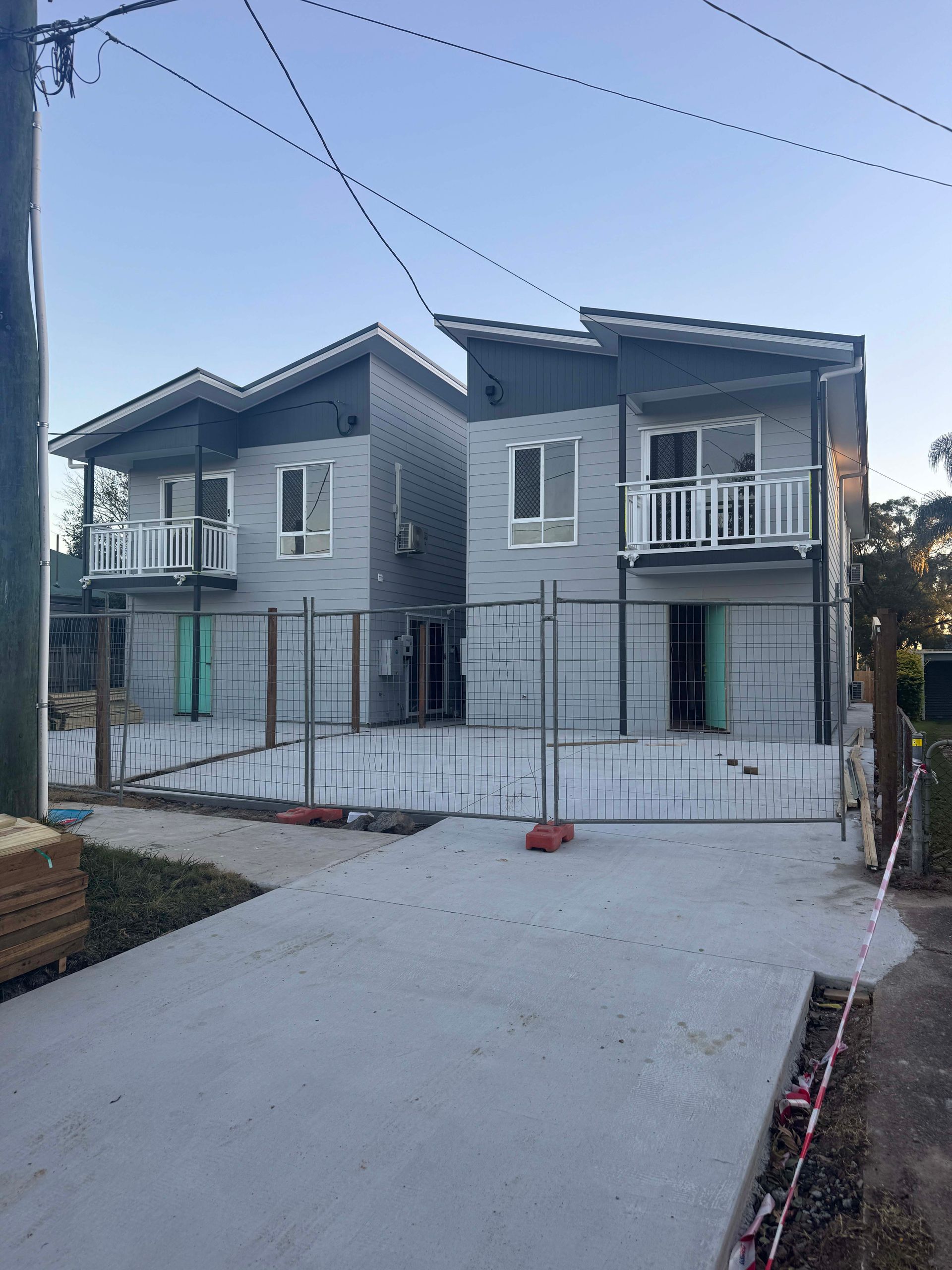 Two-story gray duplex under construction, each with balcony and white railings, set on a concrete slab, with safety fencing.
