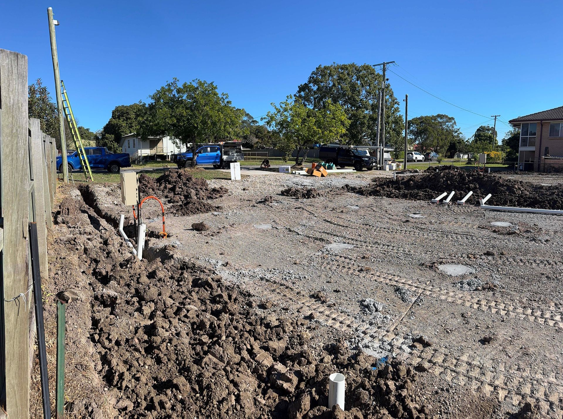 Construction site with freshly dug earth, plumbing pipes, and a blue sky. A fence is in the foreground.