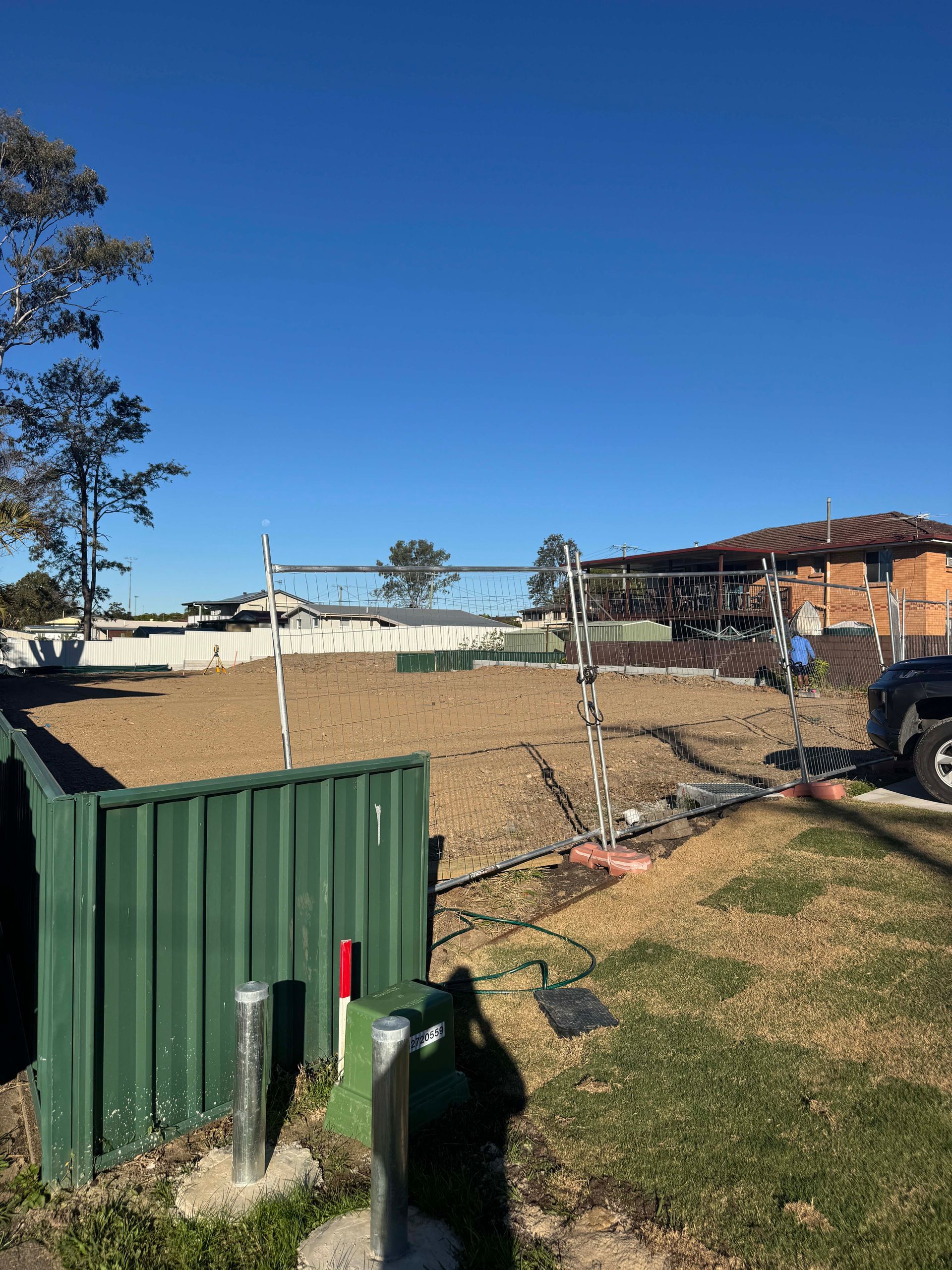 Open field with a green fence in the foreground. Construction scaffolding and a brick house are visible in the distance.
