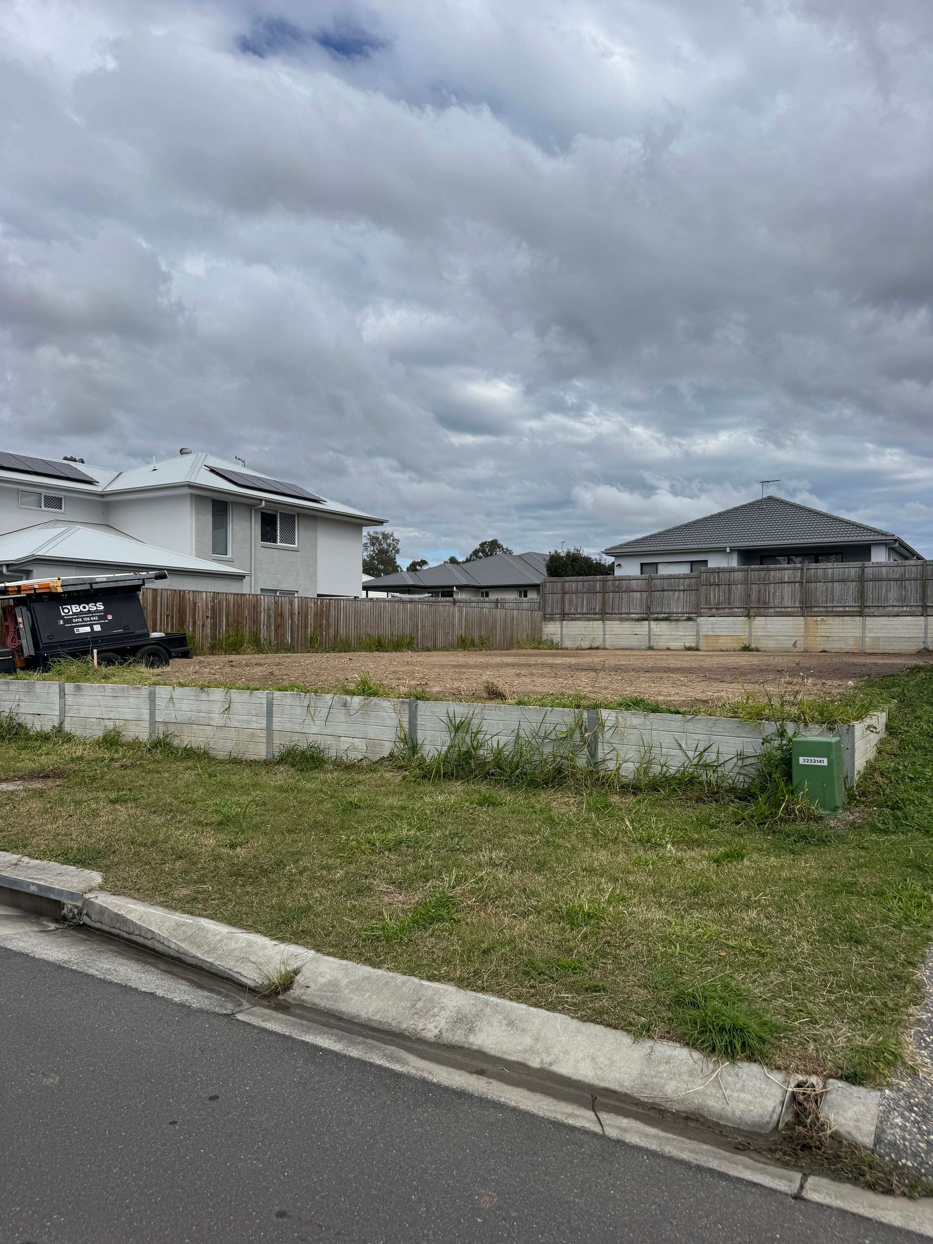 Empty building lot behind a concrete wall and curb, with houses visible in the background under a cloudy sky.