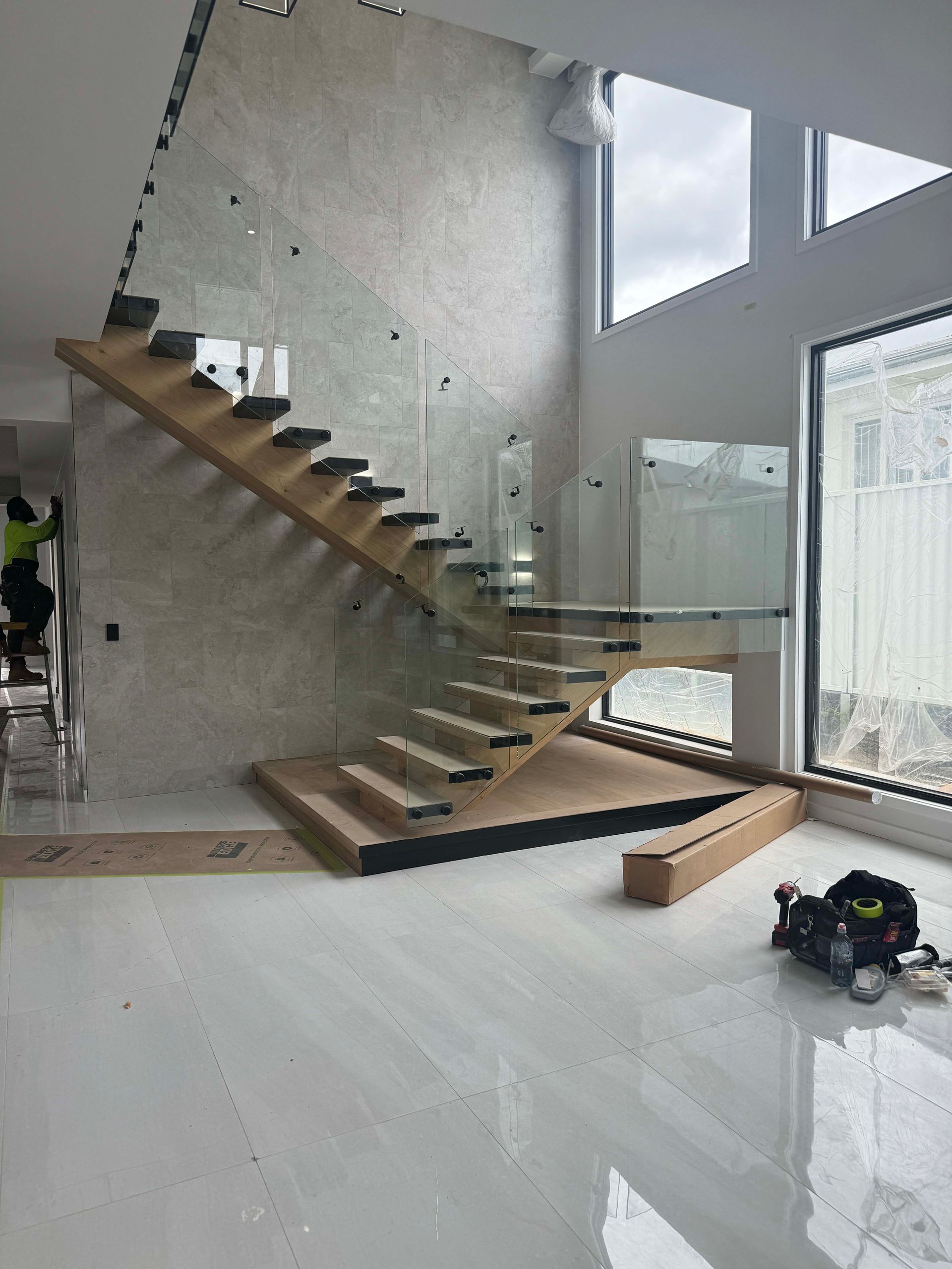 Floating wooden staircase with glass railing in a modern home. Light wood steps, white walls, and large windows are featured.