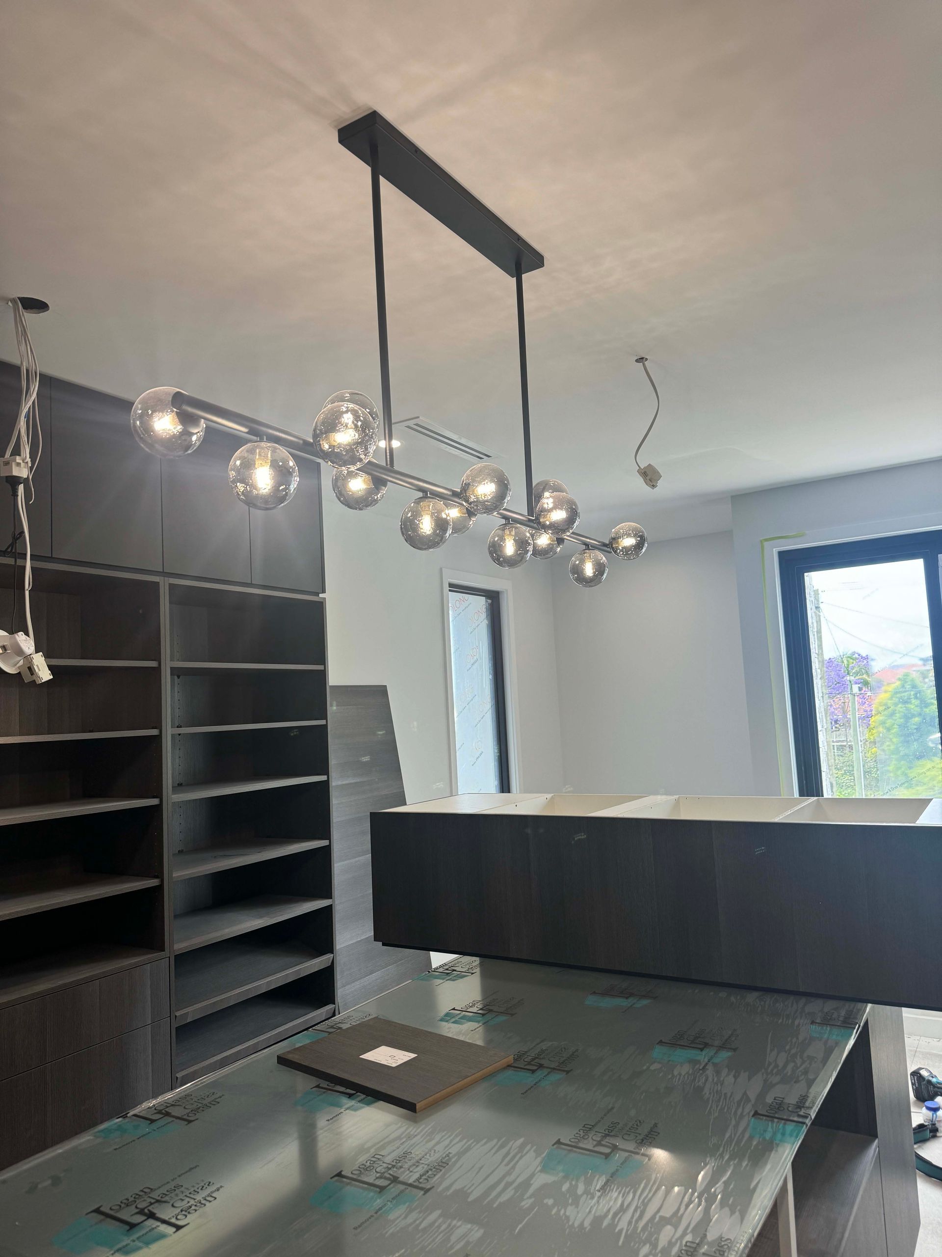 A modern chandelier with spherical lights hangs above a dark wooden counter and shelving in a room with a window.